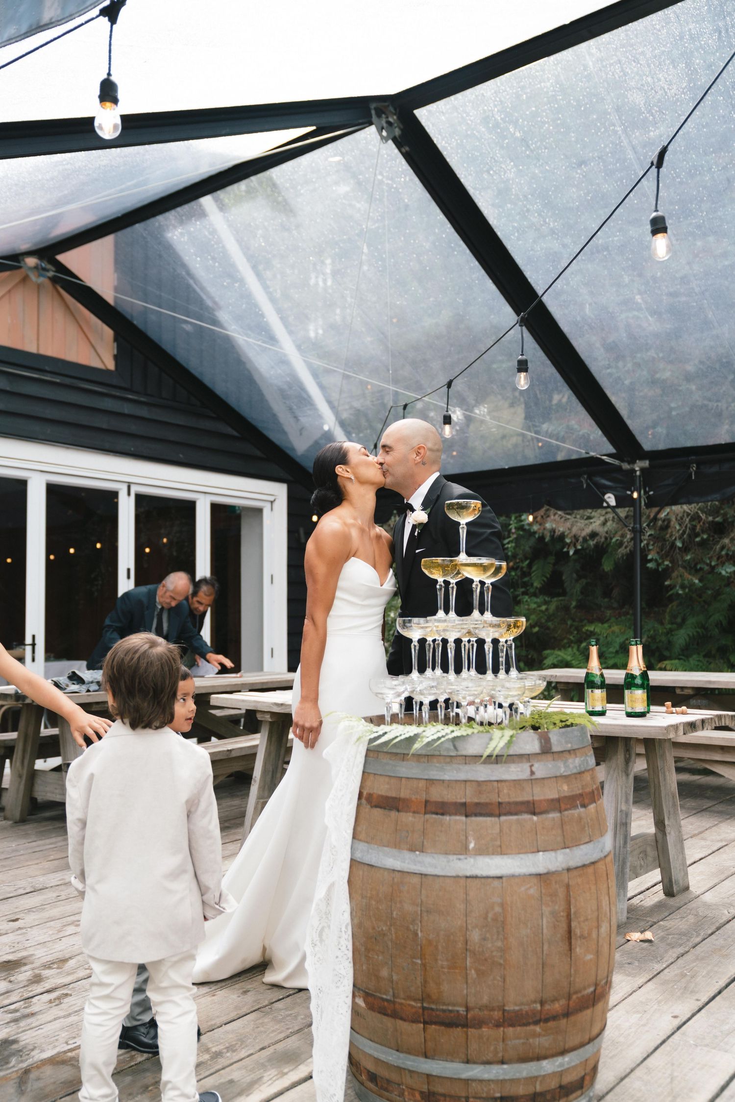 Newlyweds pose beside a champagne tower on a wooden barrel deck under a clear tent with string lights.