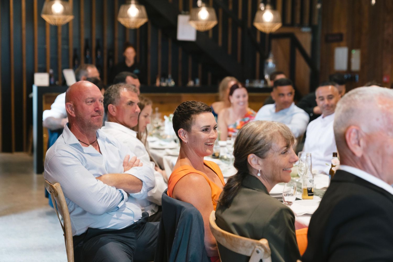 Guests seated at long tables enjoy a social gathering in a warmly lit restaurant interior.