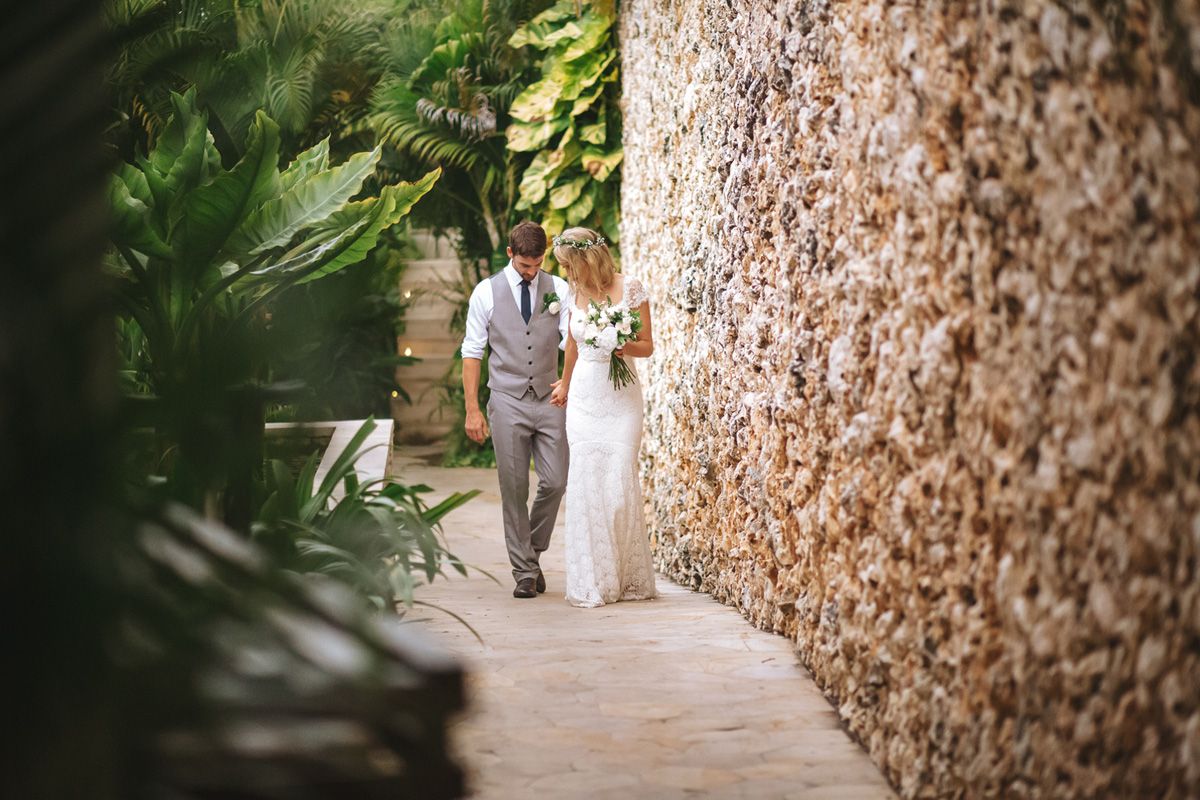 A wedding couple walks along a stone-walled pathway with tropical foliage.