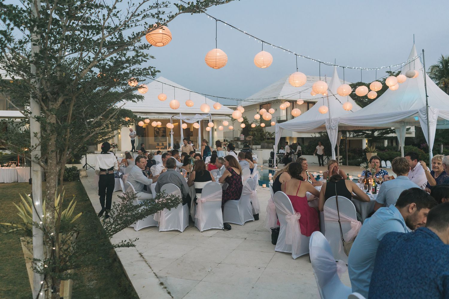 Outdoor wedding reception under string lights with white tents and tables at dusk.