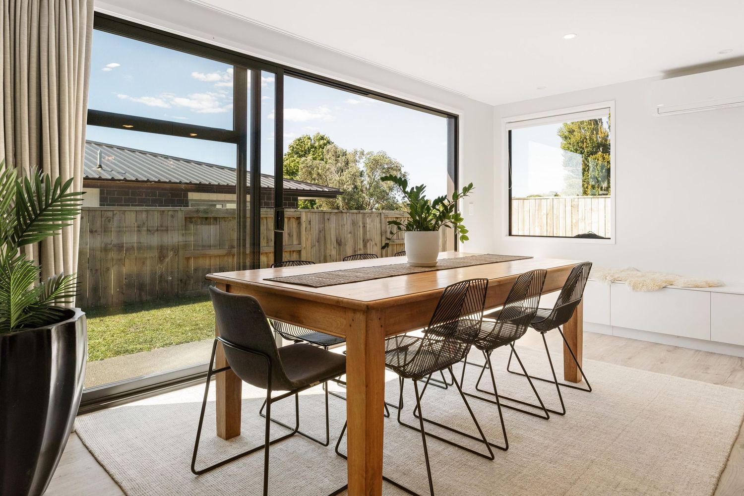 Modern dining area with wooden table, black chairs and large sliding glass doors opening to outdoor patio space.
