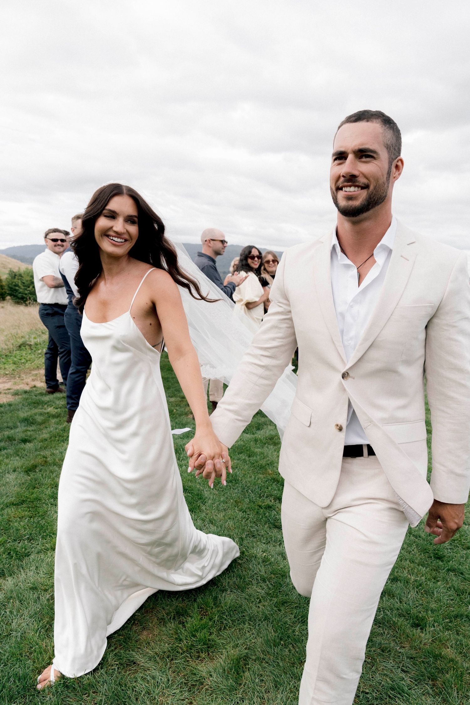 A happy couple in white attire running joyfully across a grassy field during their wedding celebration.