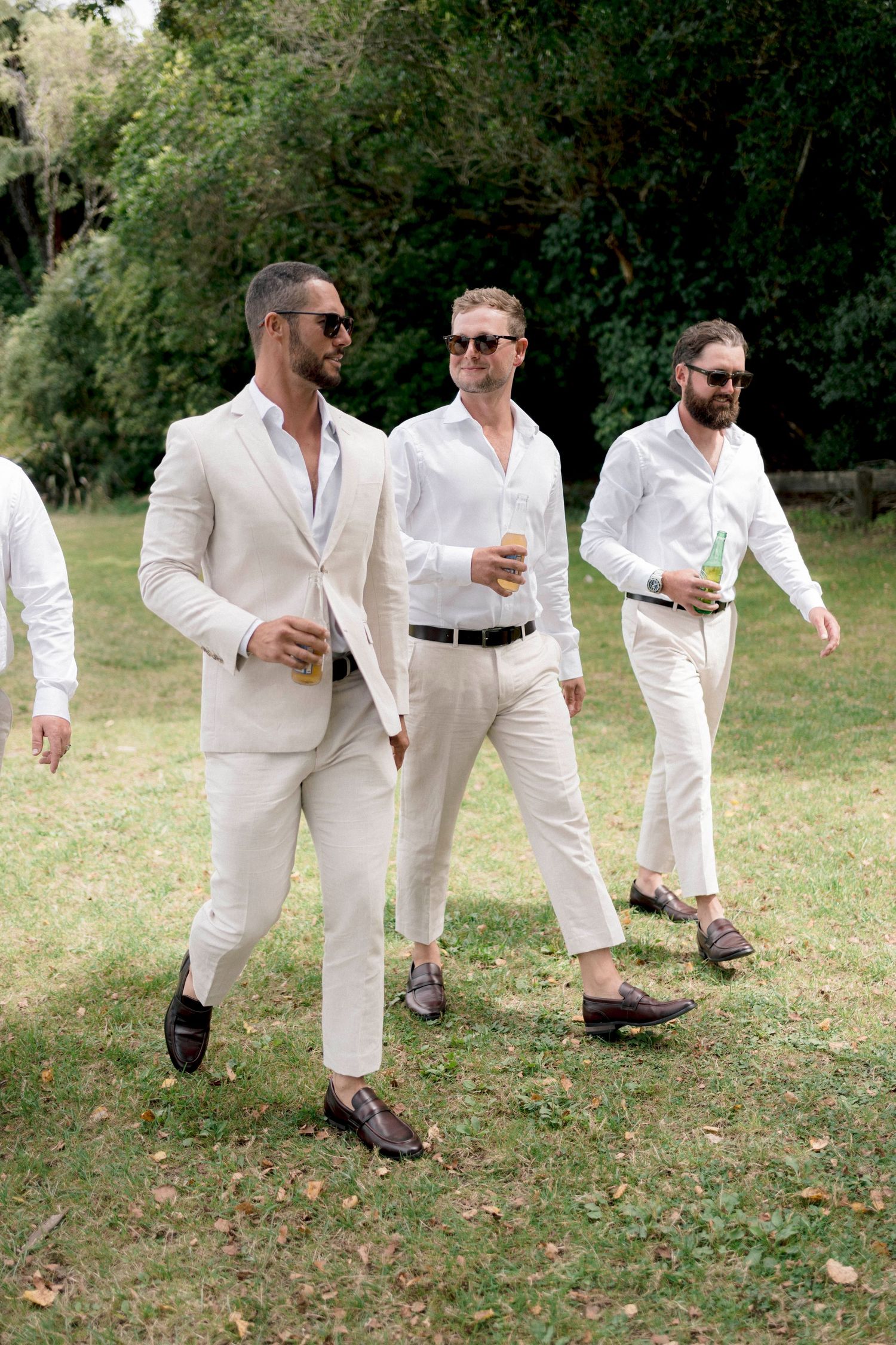 Groomsmen in light colored suits walking and socializing outdoors on grass.