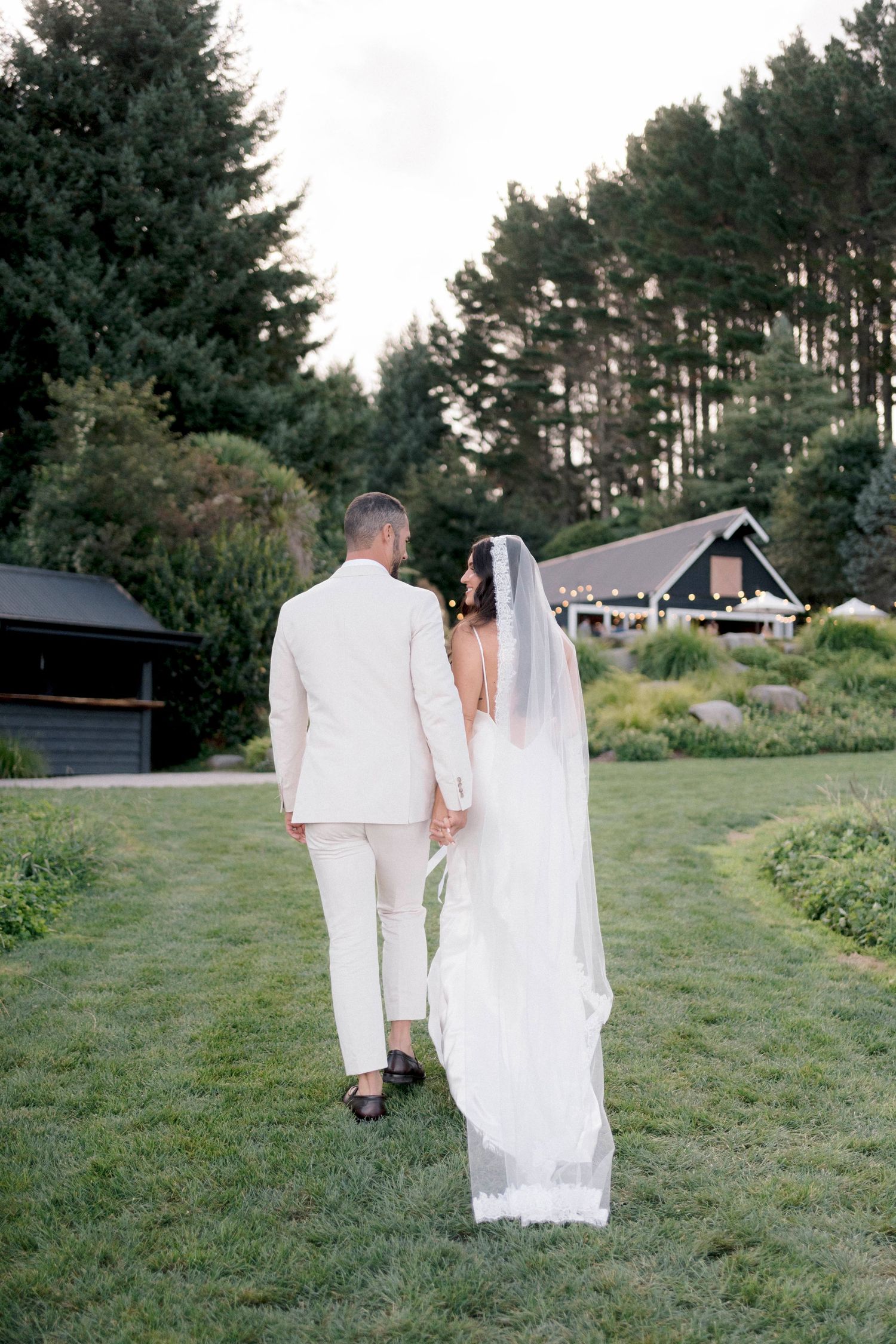 A wedding couple in white attire stands on a lawn with evergreen trees and a rustic building in the background.