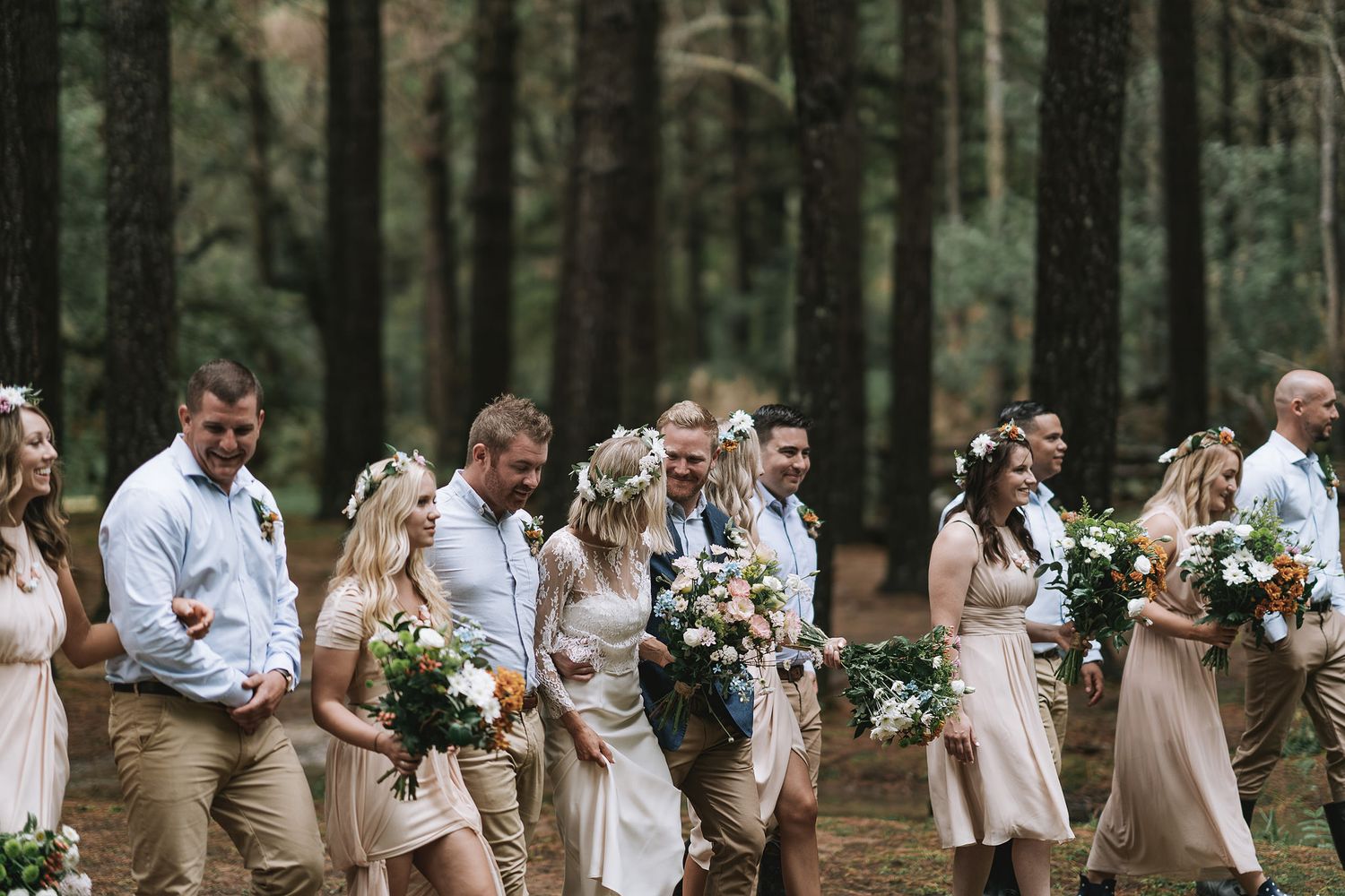 Wedding party in casual attire stands together in a scenic forest setting holding floral bouquets.