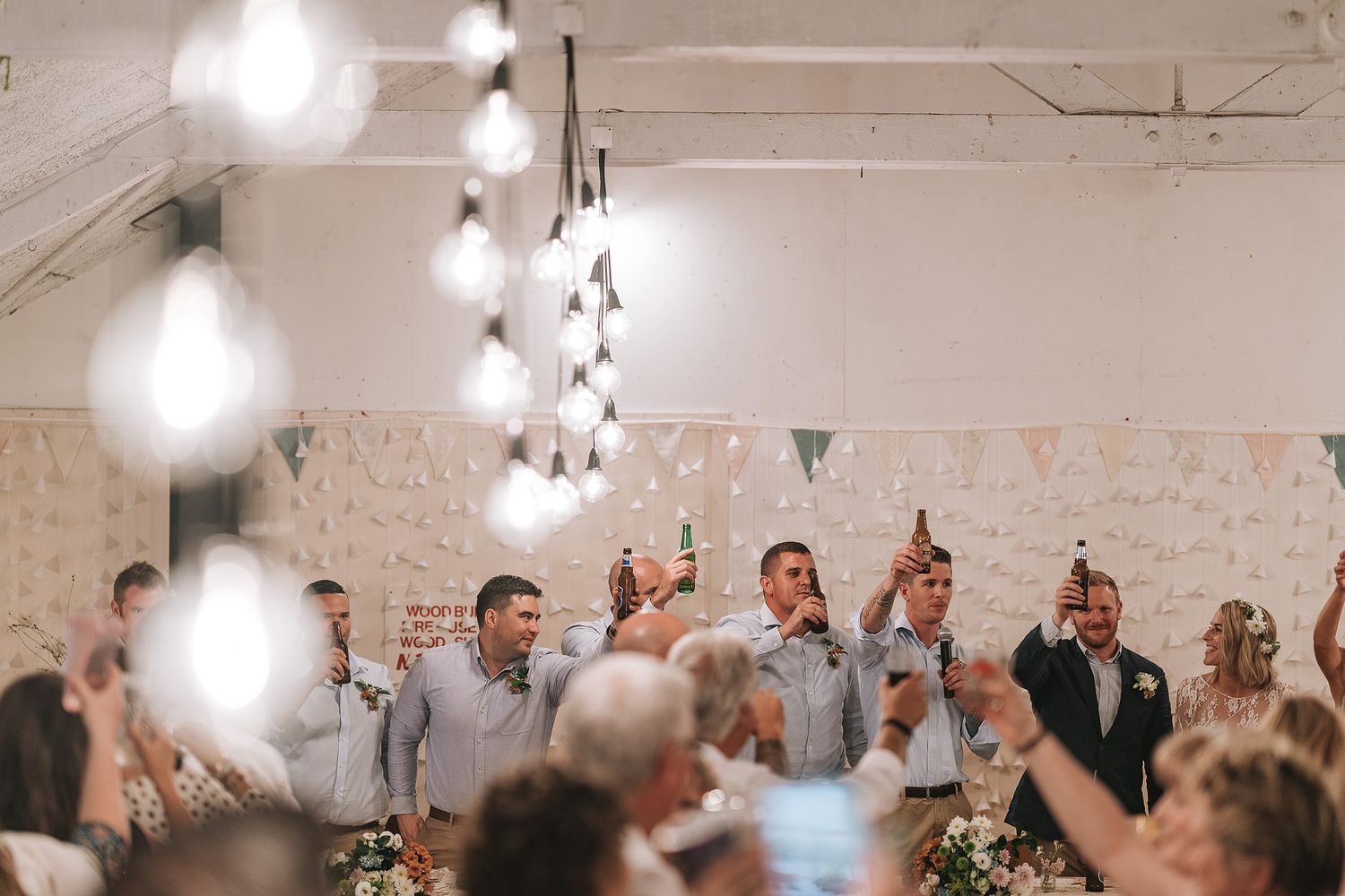 Wedding reception guests celebrate and raise their glasses for a toast in a brightly lit venue with hanging string lights.