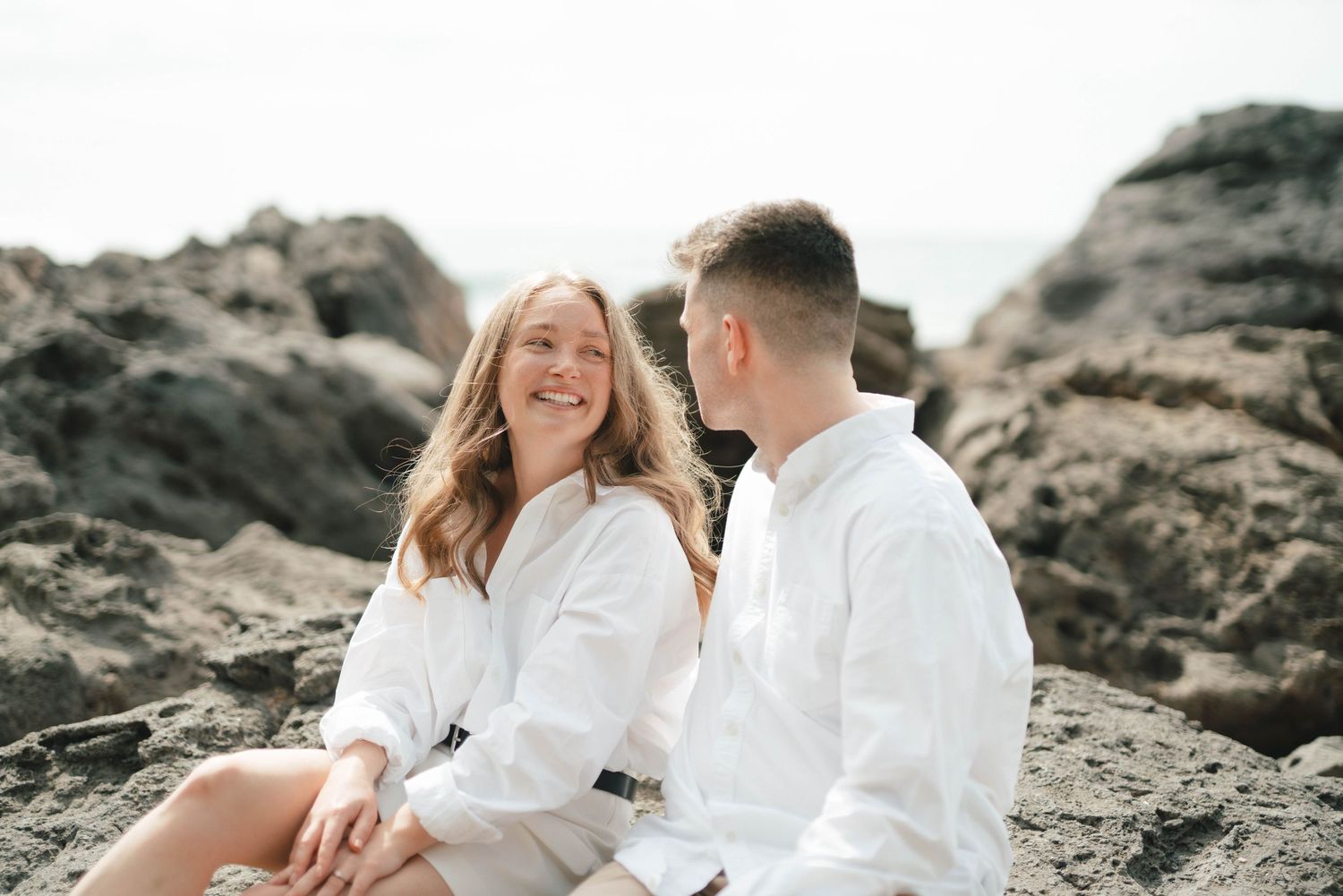A couple dressed in white embraces on a rocky beach with crashing waves and dramatic coastline in the background.