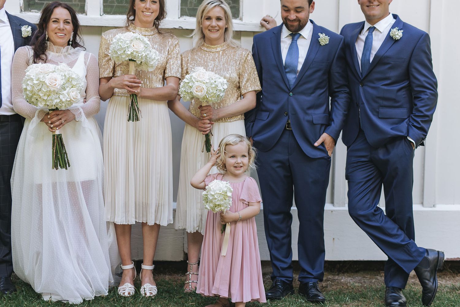 Wedding party group including bridesmaids in blush dresses and groomsmen in navy suits standing together outside.