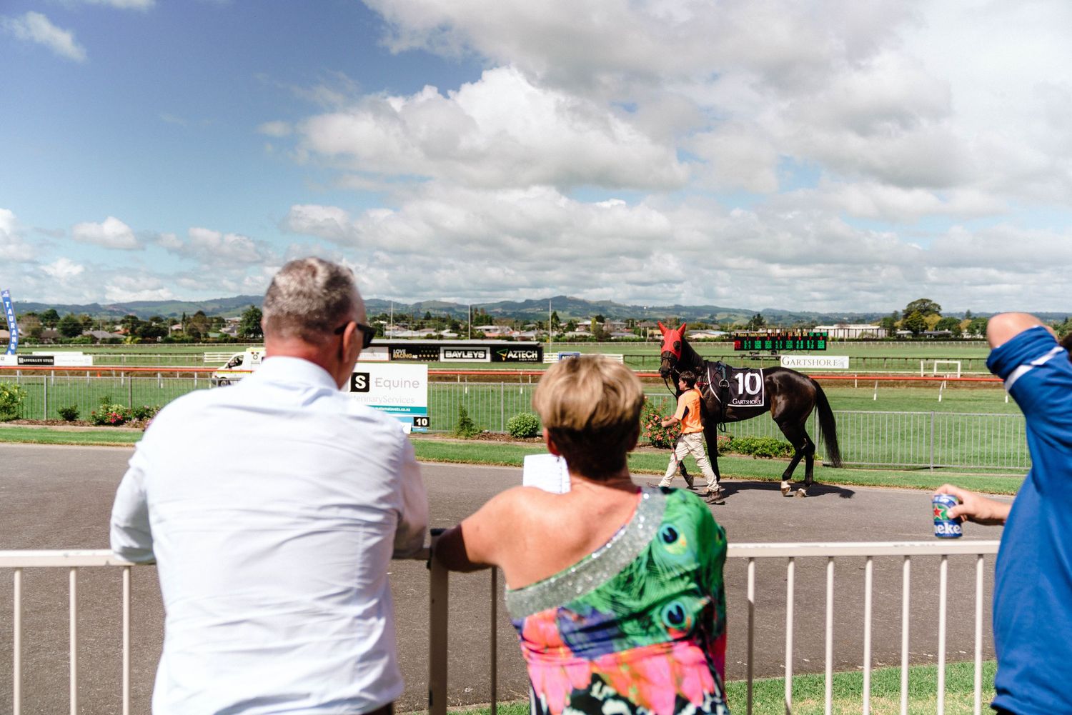 Spectators watch from the railing at an outdoor equestrian event on a sunny day.