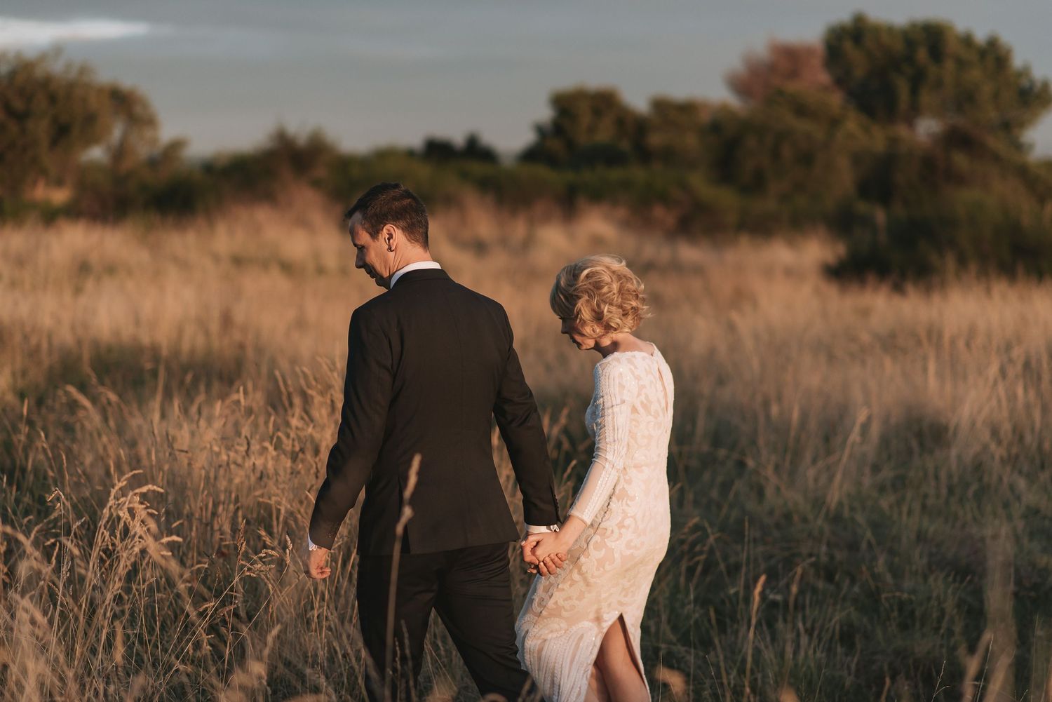 Couple in elegant attire walking hand in hand through golden tall grass field at sunset.