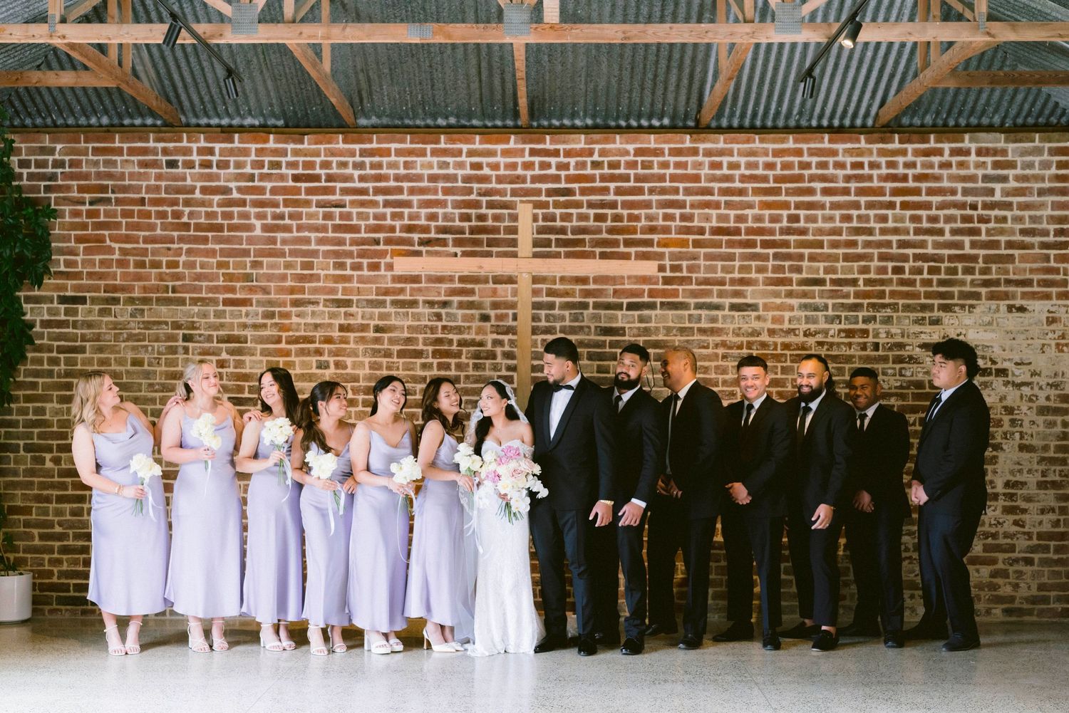 Wedding party lined up against rustic brick wall wearing formal black and white attire.