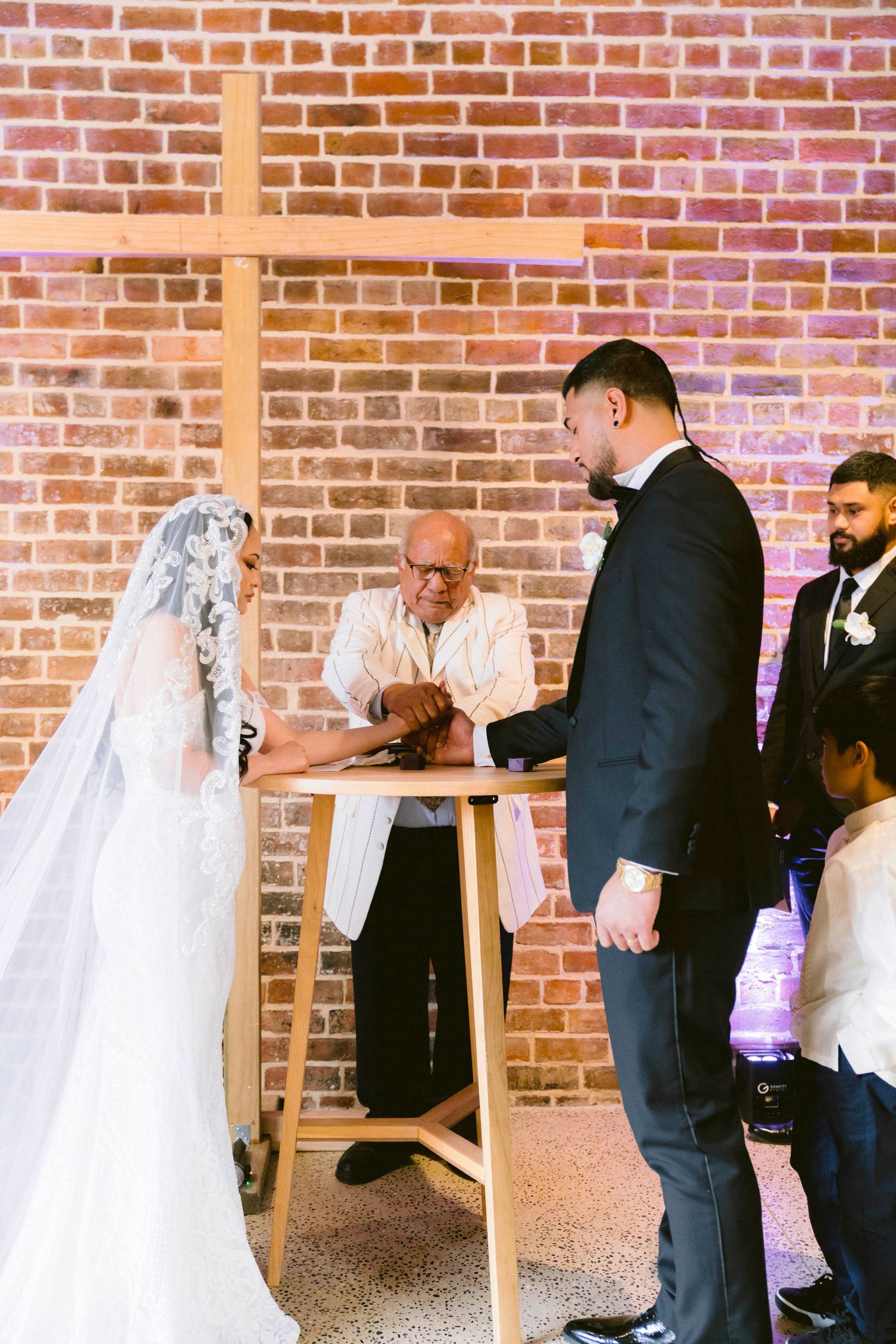 A wedding ceremony taking place in front of a wooden cross and exposed brick wall with purple lighting.
