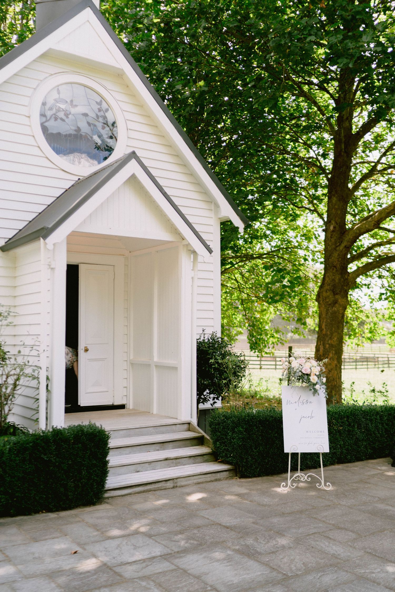 Charming white wedding chapel nestled among lush green trees with paved walkway leading to entrance.
