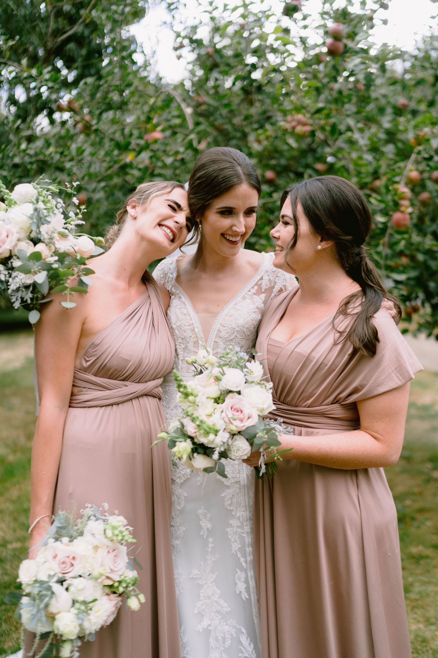 Three women share joyful laughter among apple trees while holding wedding bouquets.