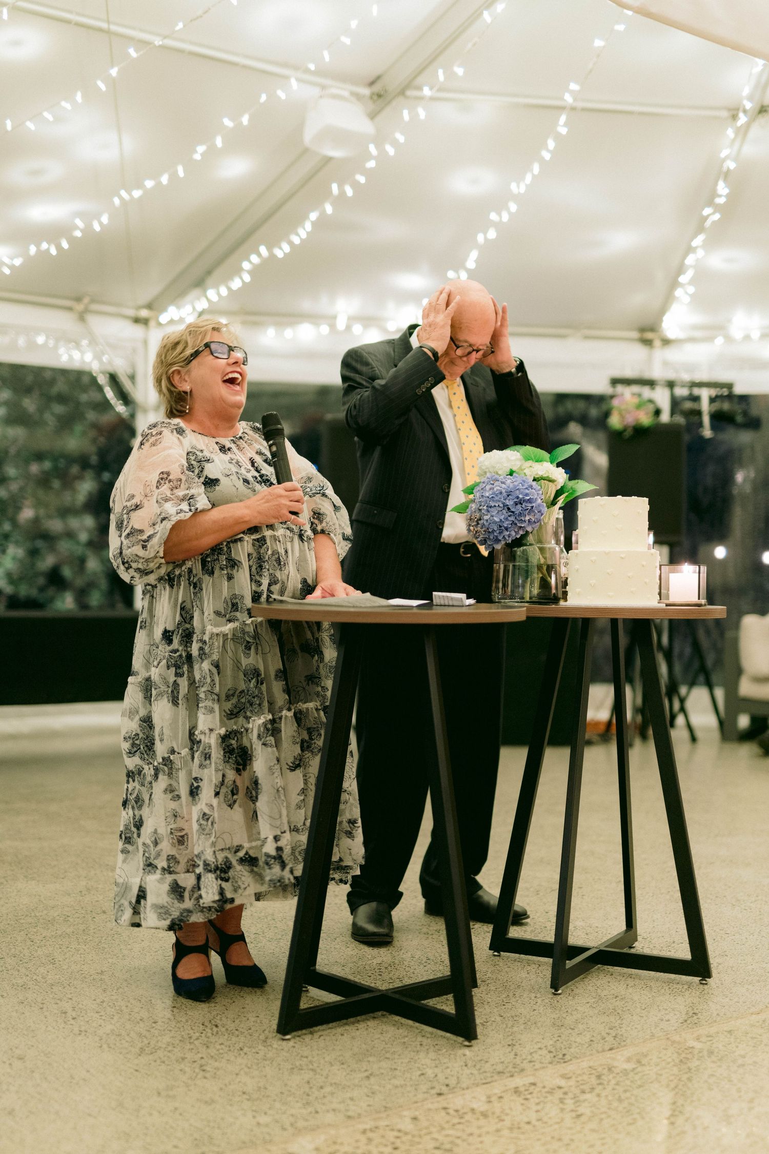 Wedding guests share laughter during cake cutting ceremony under a white tent decorated with twinkling string lights.