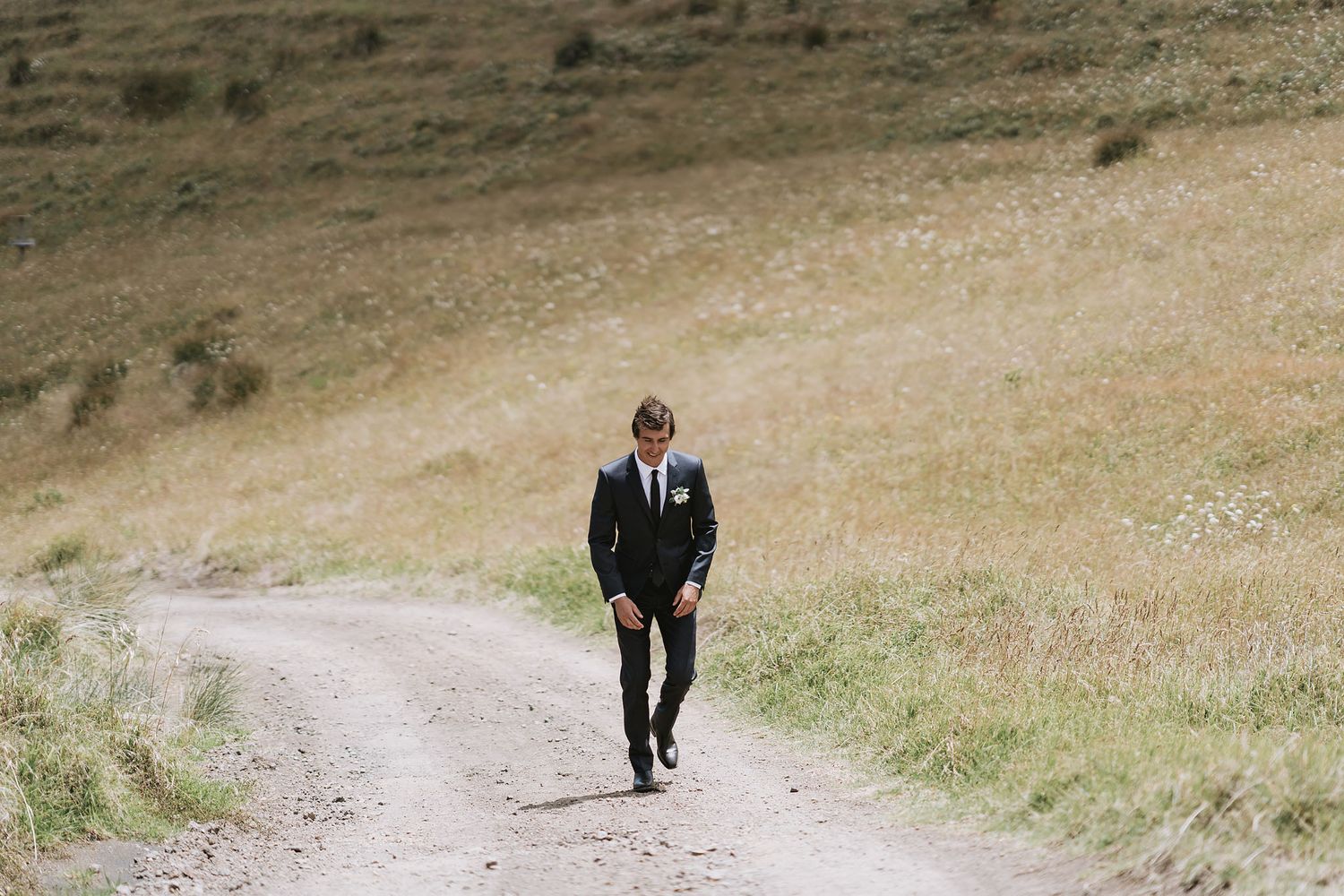 A person in a black suit walks alone on a winding dirt path through rolling hills on an overcast day.