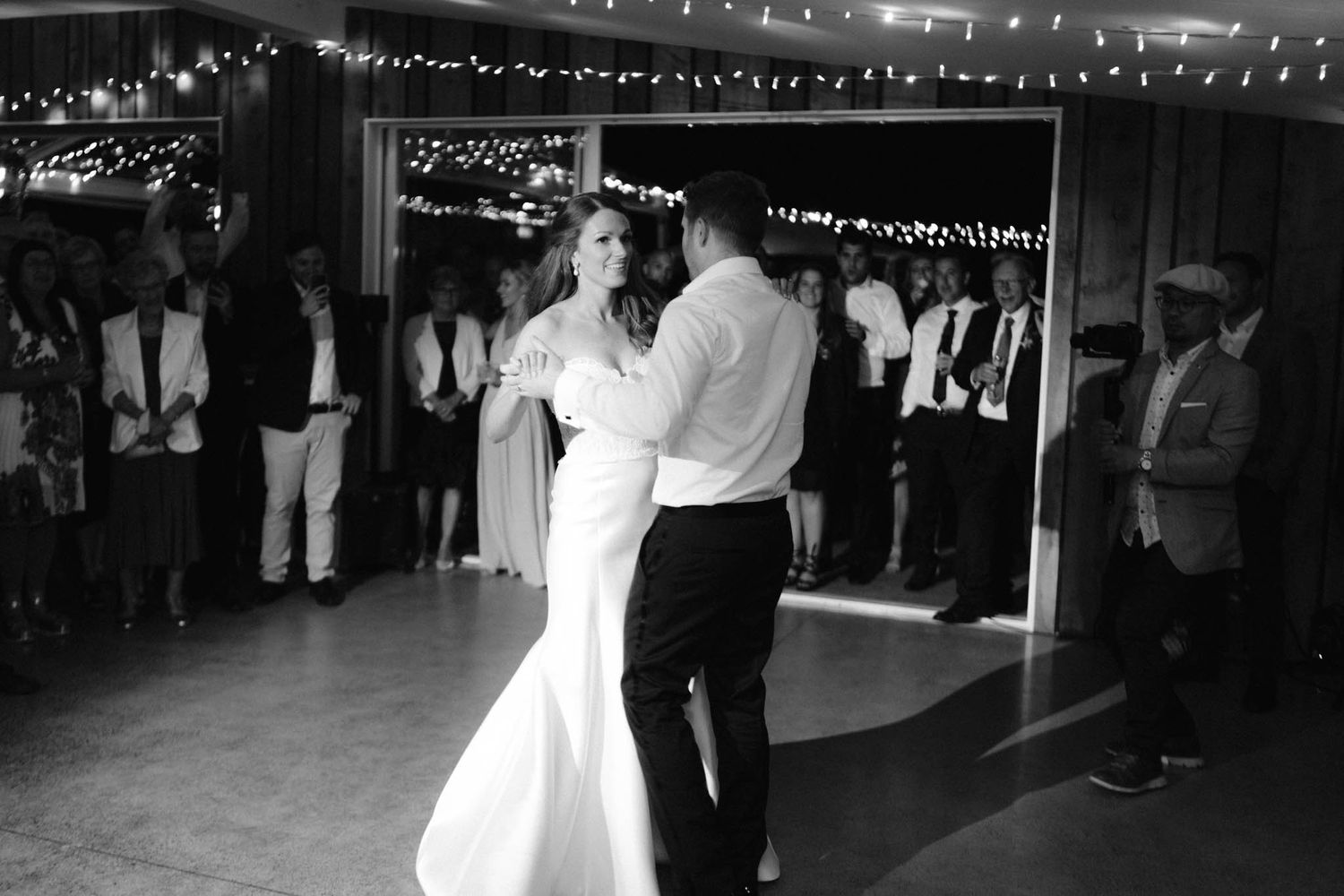 A couple shares their first dance as newlyweds under twinkling lights at their wedding reception in black and white.