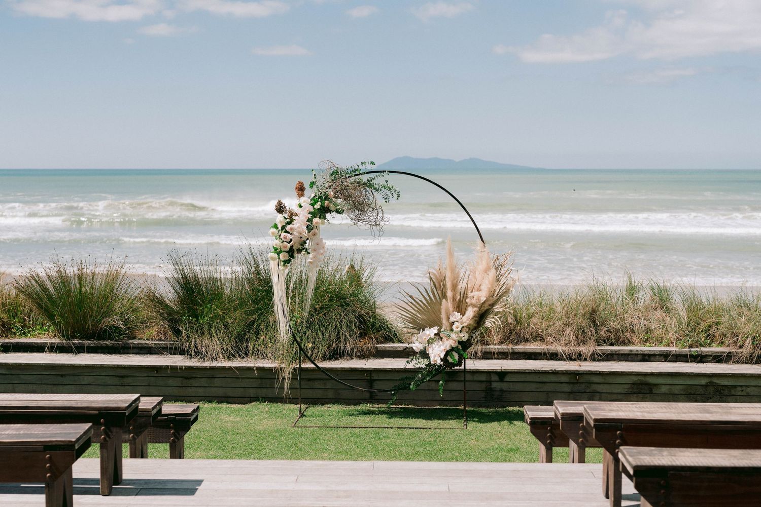 Coastal wedding ceremony setting with ocean views and wooden benches on a sunny beach day.