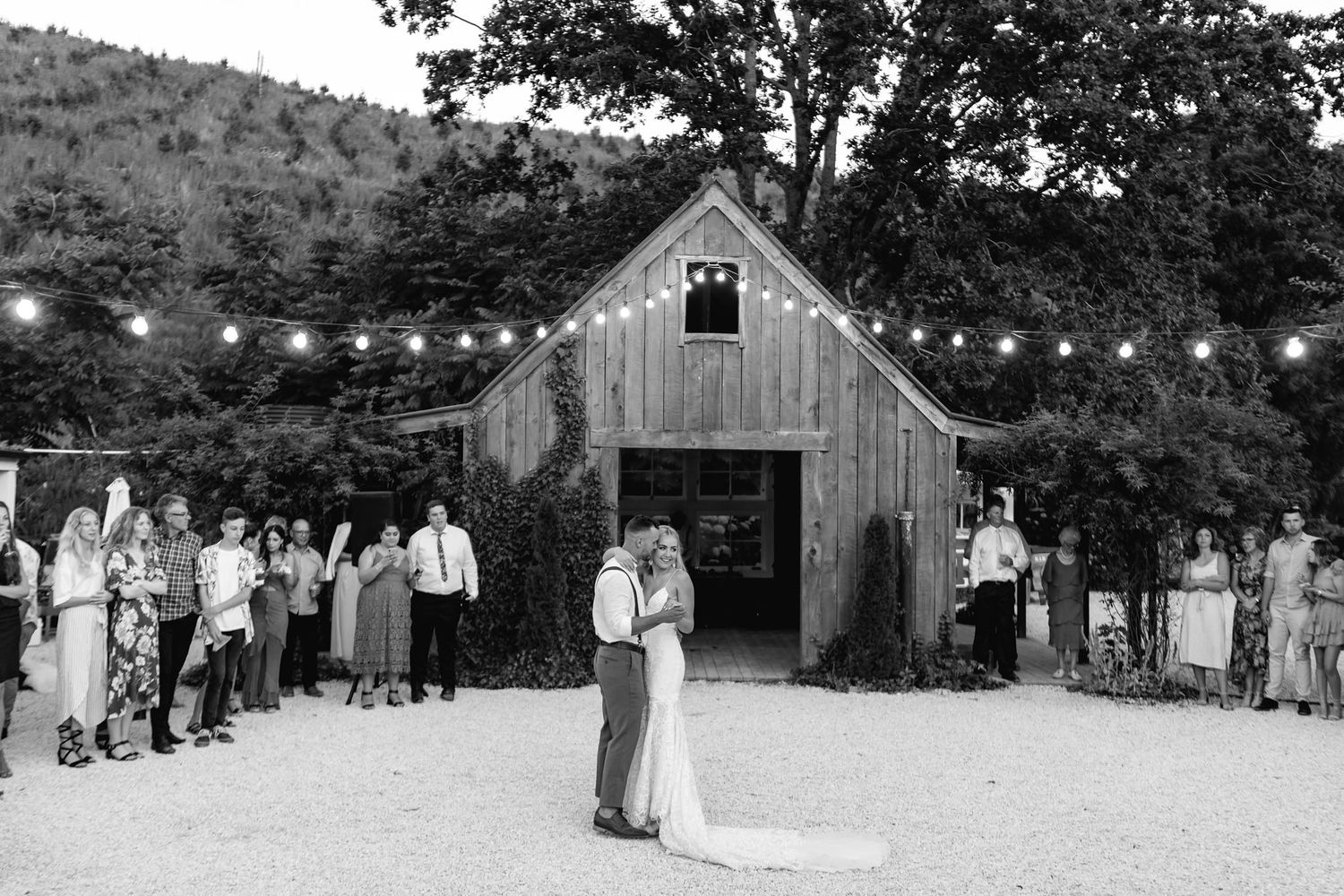 Guests gather at a rustic barn wedding venue decorated with string lights and surrounded by greenery for an evening celebration.