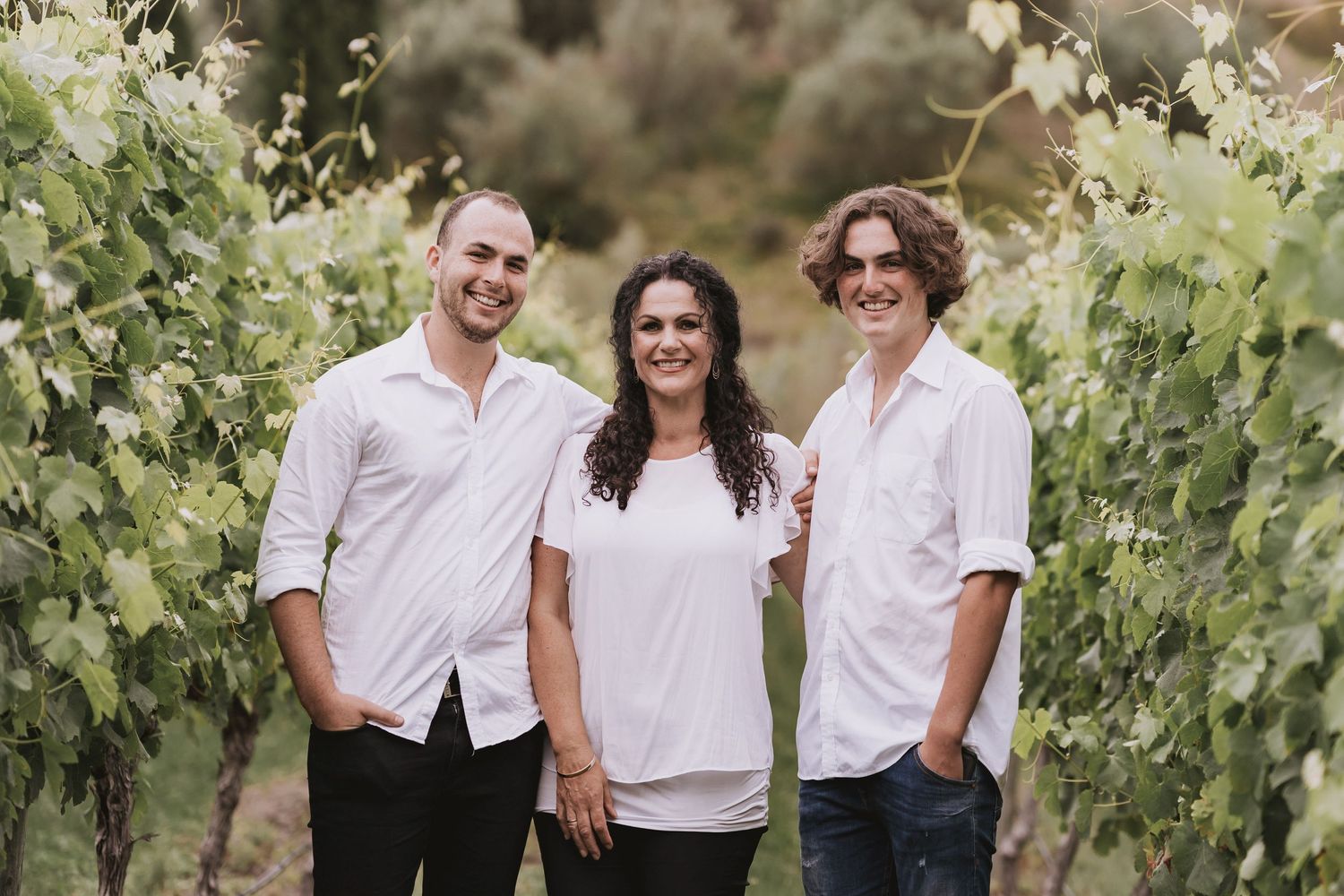 Three people in white shirts smiling while standing together in a vineyard on a sunny day.
