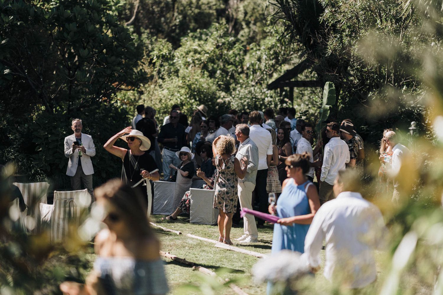 A group of guests gathers outdoors for a casual daytime event in a lush garden setting.