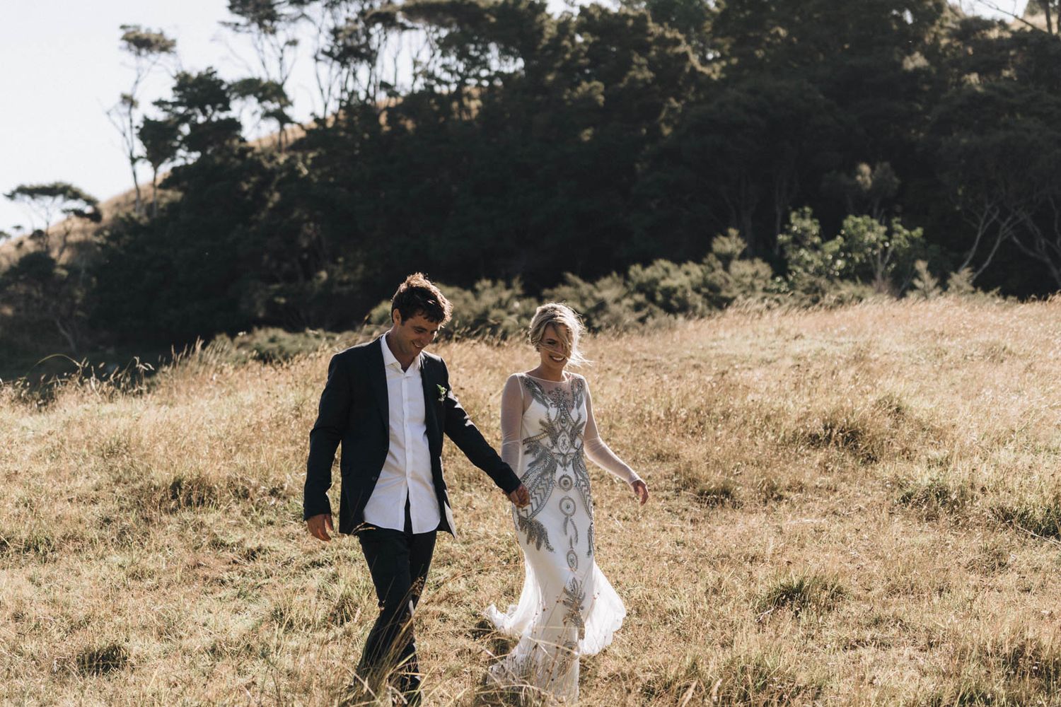 A newlywed couple walks hand in hand through a sunlit meadow with pine trees in the distance.