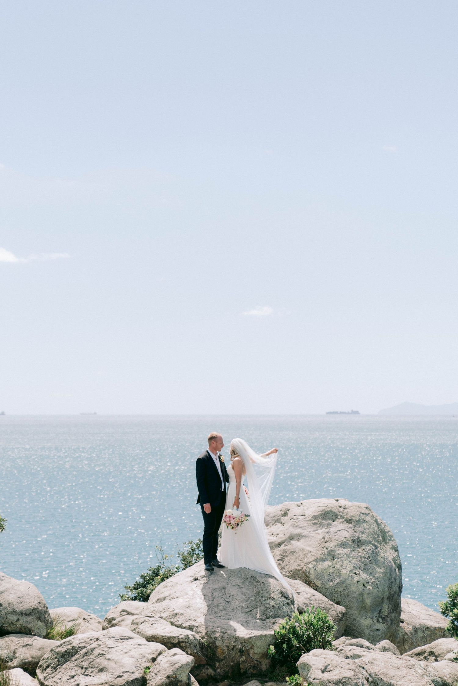 A couple poses on rocky coastal cliffs overlooking a sparkling ocean on their wedding day.