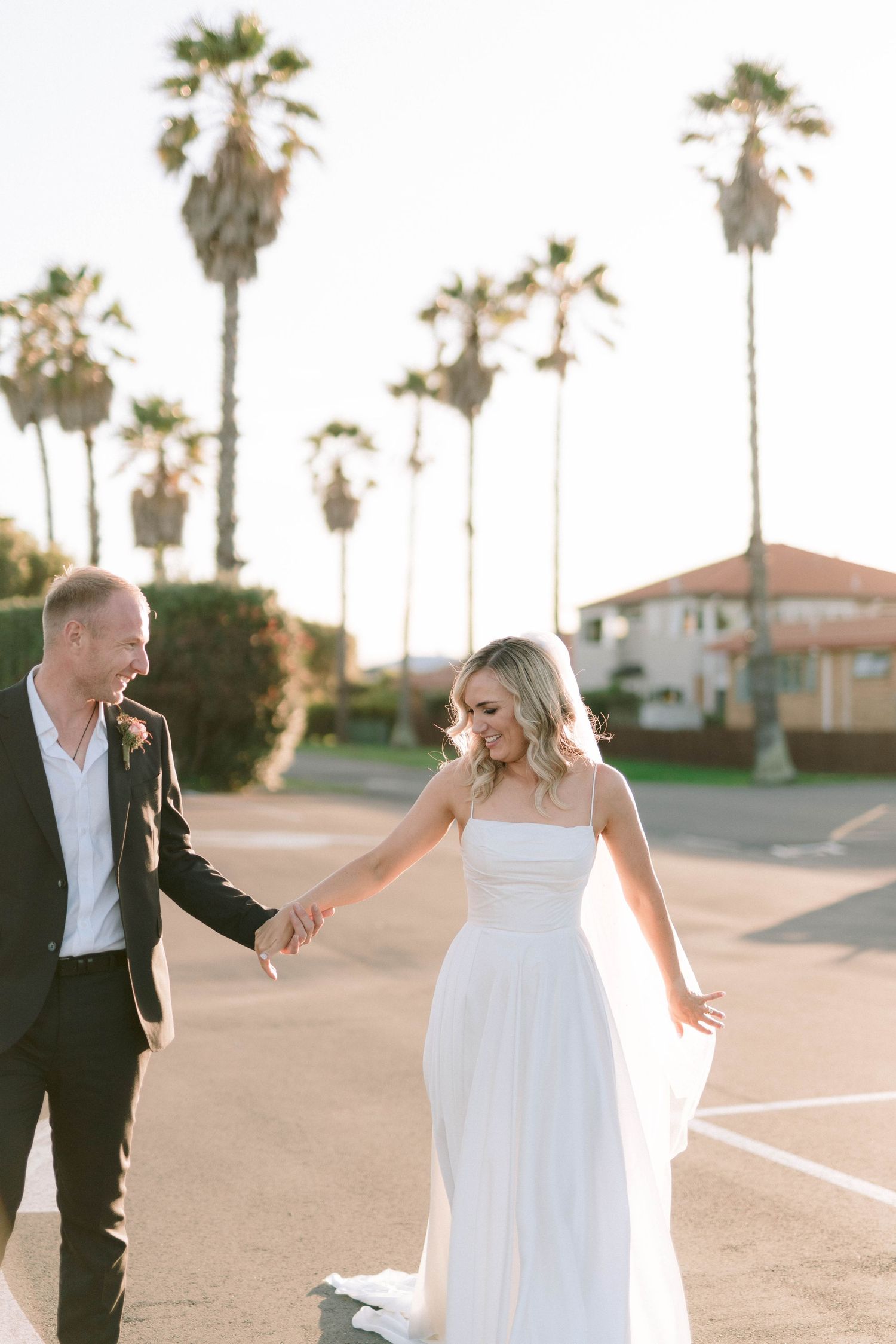 Bride in white gown and groom in black suit dance playfully on a palm tree-lined path at sunset.