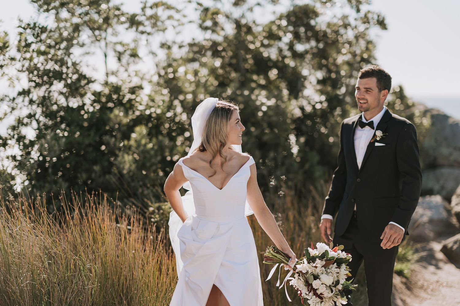 Couple in formal wedding attire standing together in golden field during sunset with white floral bouquet.