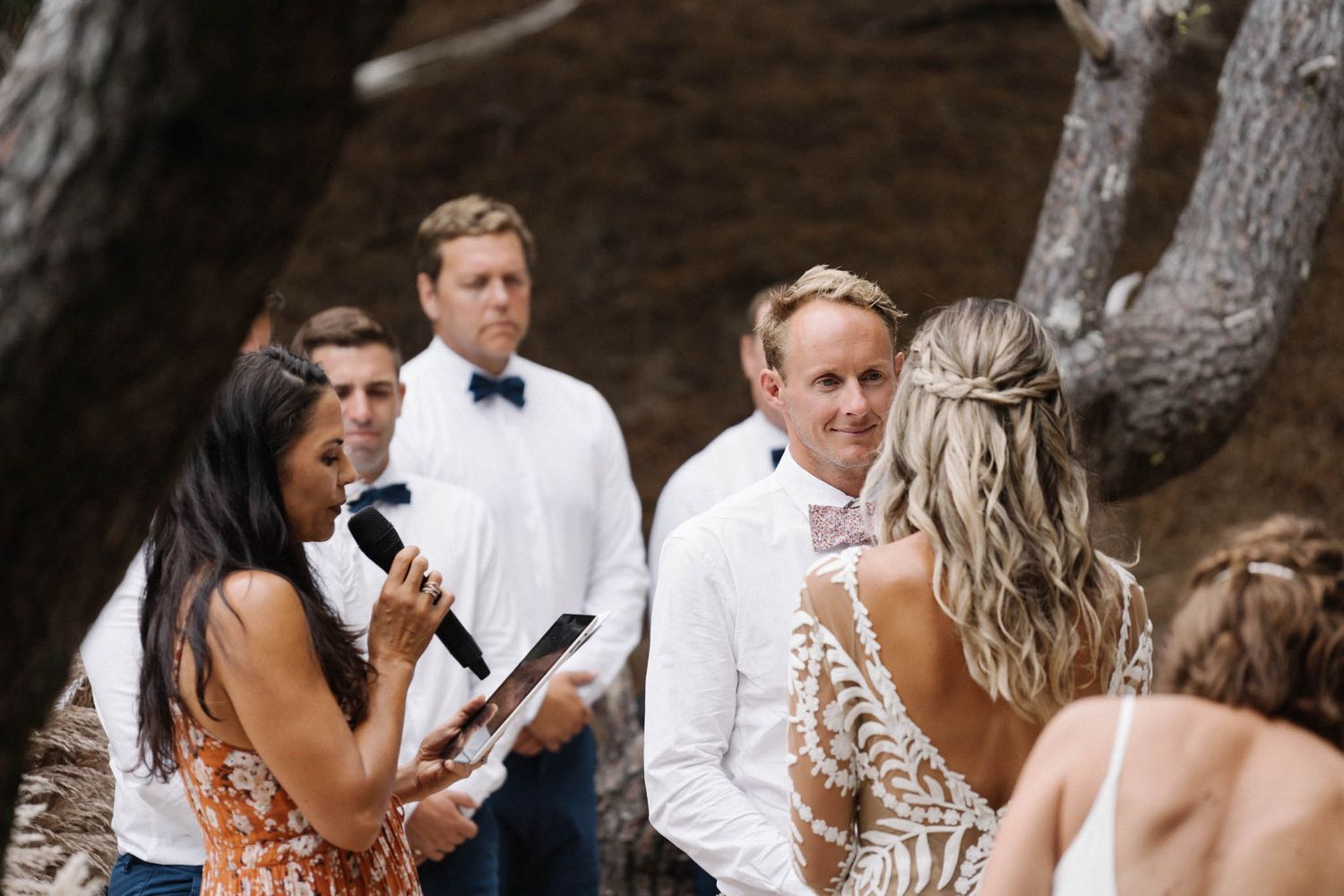 Outdoor wedding ceremony showing guests in formal attire gathered under large tree branches.