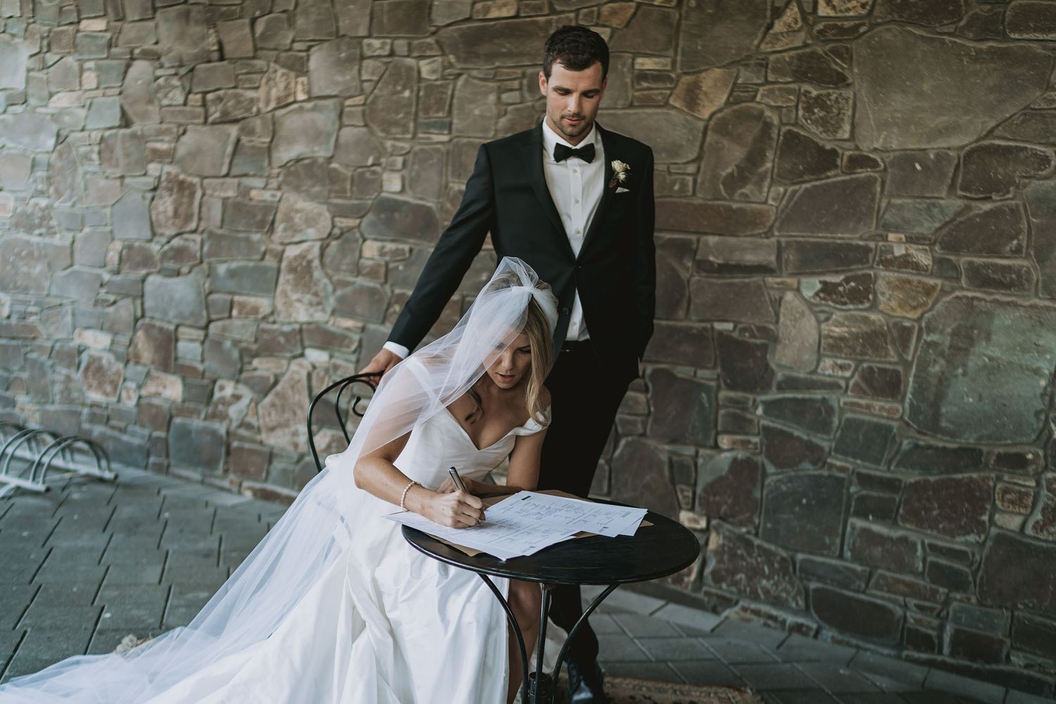 A bride in a white wedding dress signs papers while standing with a groom in a tuxedo against a stone wall.