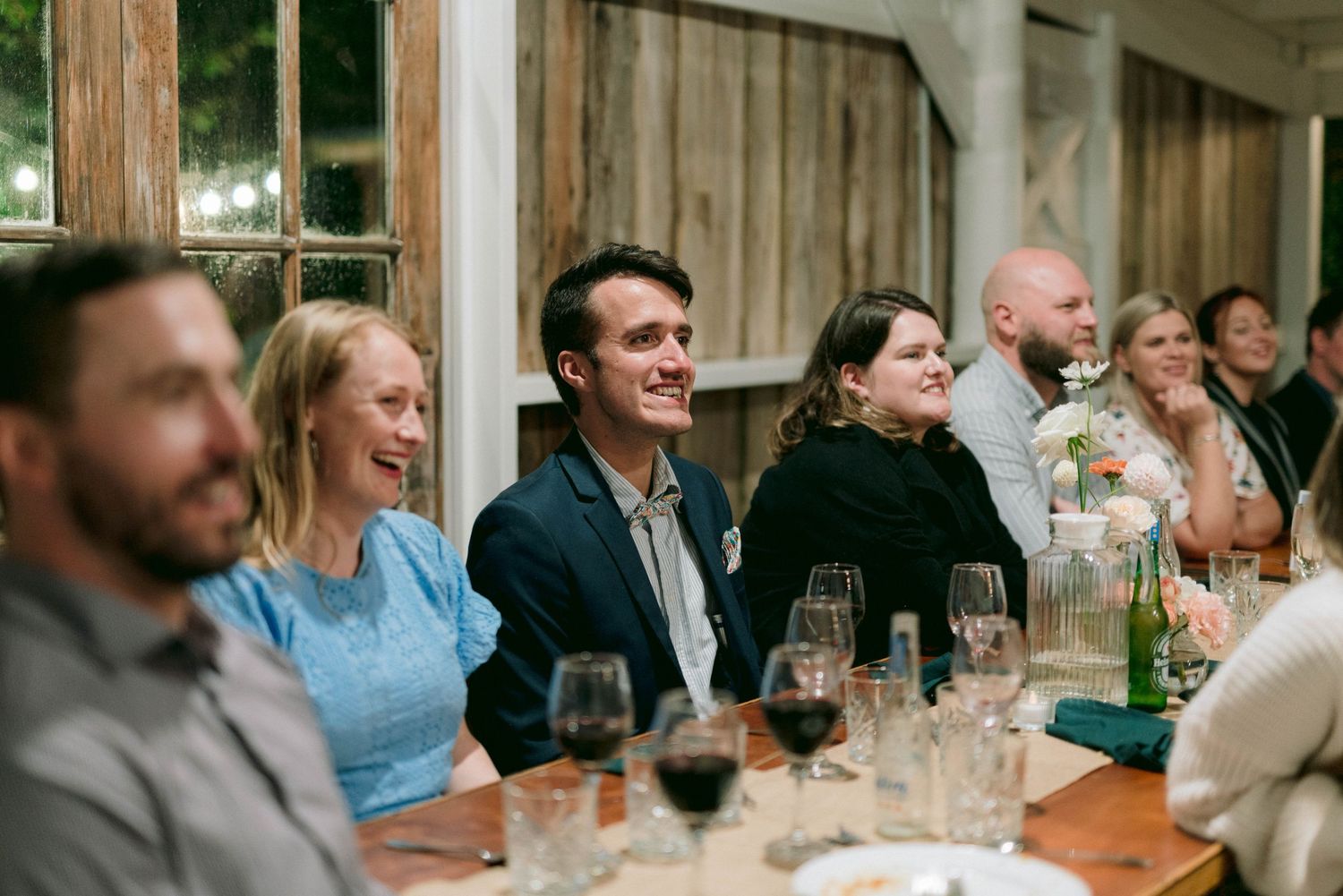 Wedding reception guests seated at long rustic wooden tables sharing conversation and laughter.