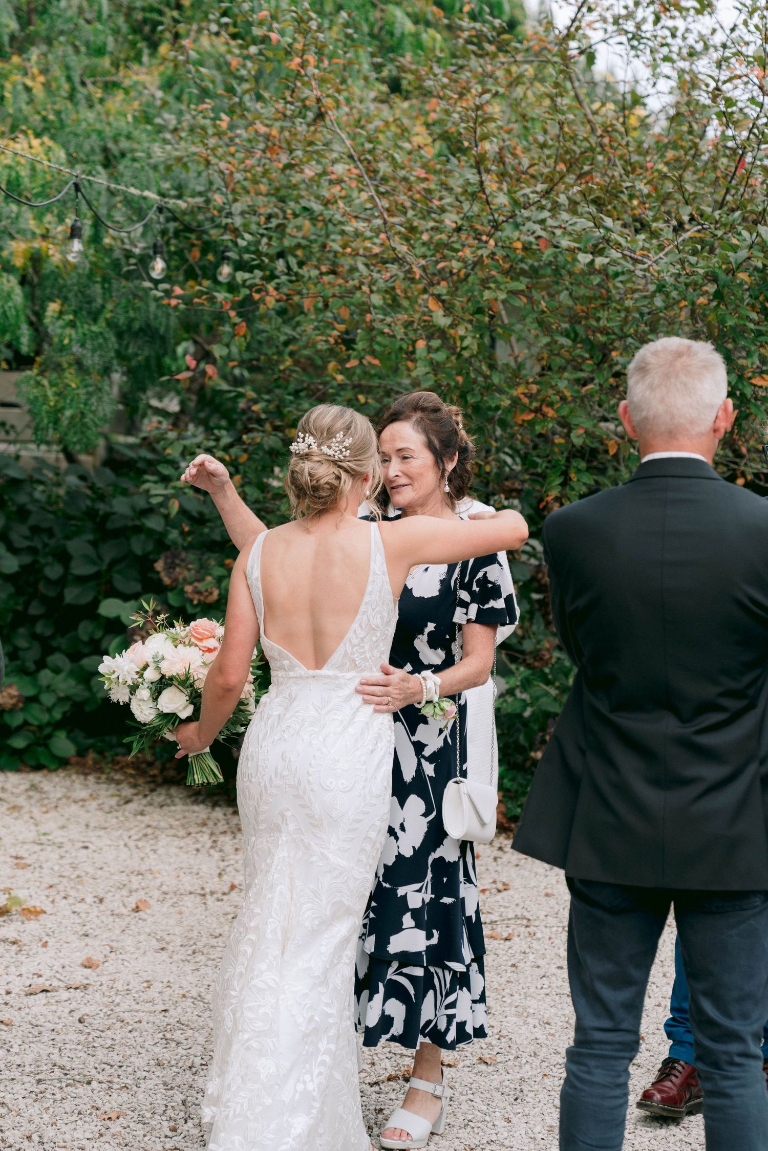 Wedding guests greet and embrace in outdoor garden setting with flowering bushes in background.
