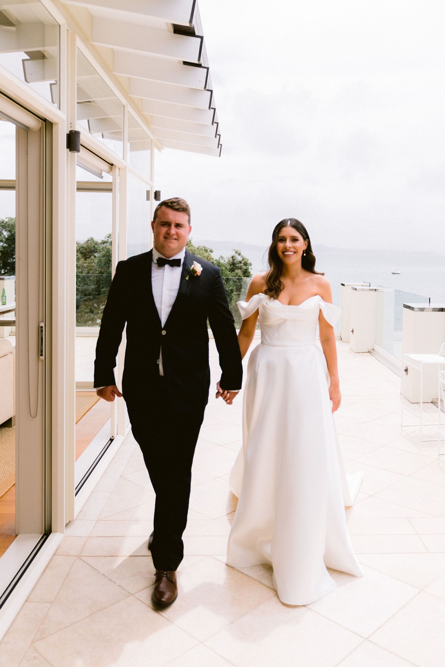 Newlyweds walk hand in hand on a bright white balcony wearing traditional wedding attire.