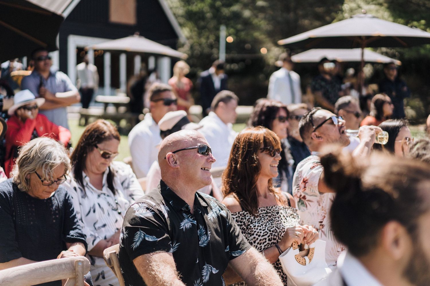 Outdoor wedding ceremony audience seated on sunny day with black barn building in background.