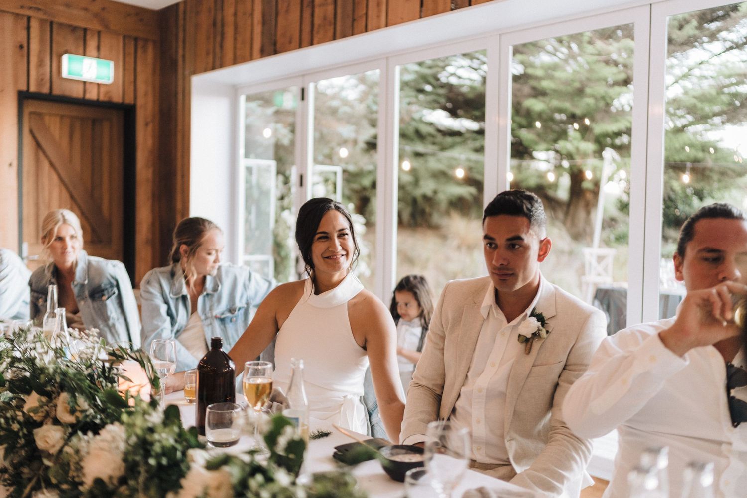 Wedding reception dinner in rustic wood-paneled room with large windows and floral centerpieces.
