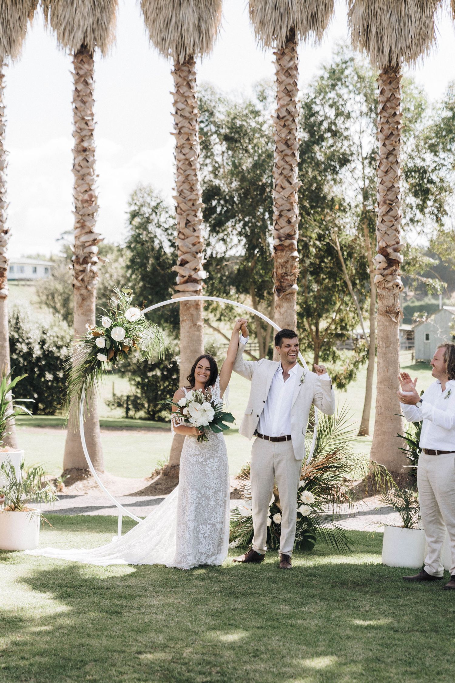 Palm trees line a grassy outdoor wedding ceremony area decorated with white floral arrangements and draping.