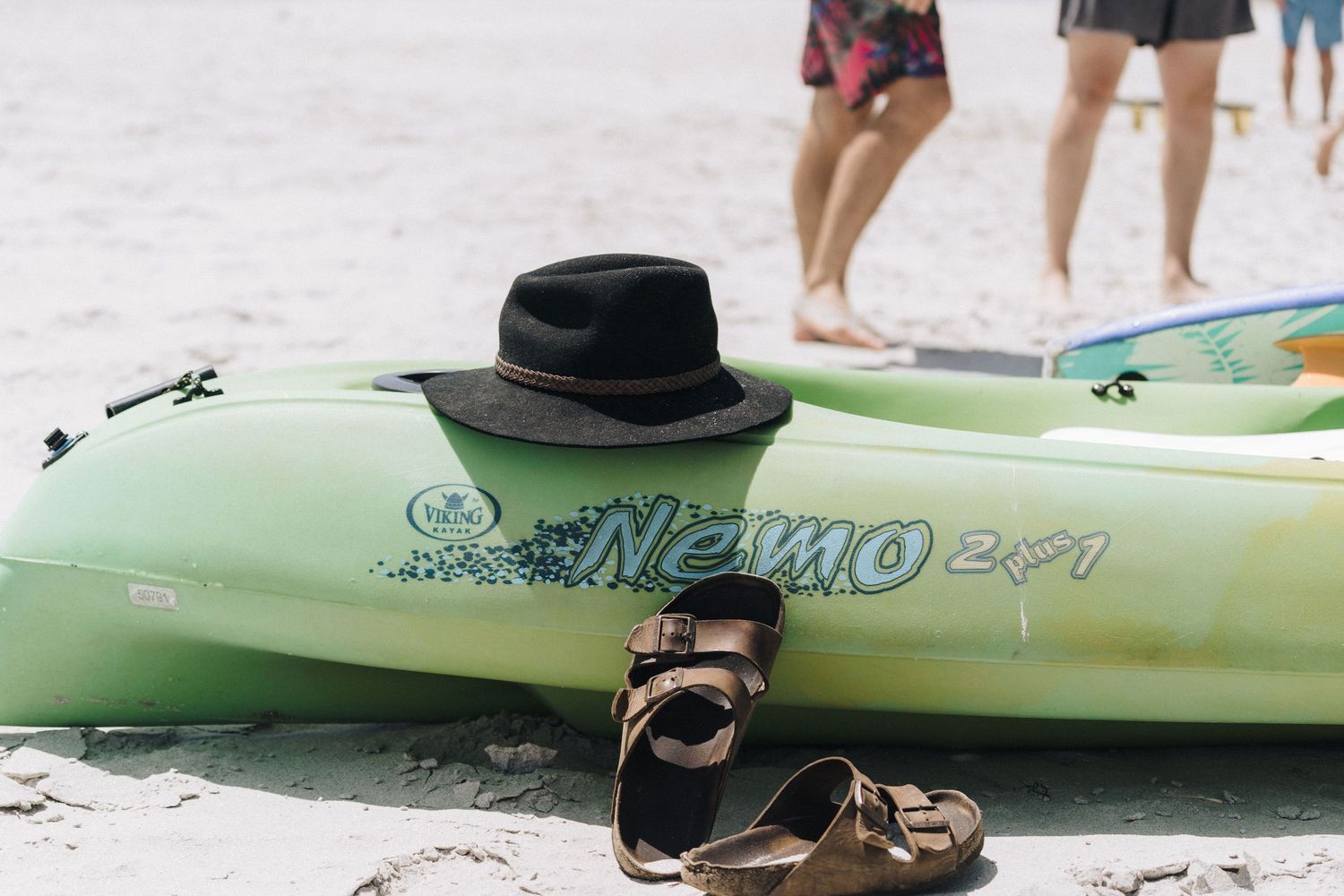 A black hat and brown sandals sit next to a light green Nemo kayak on a sandy beach.