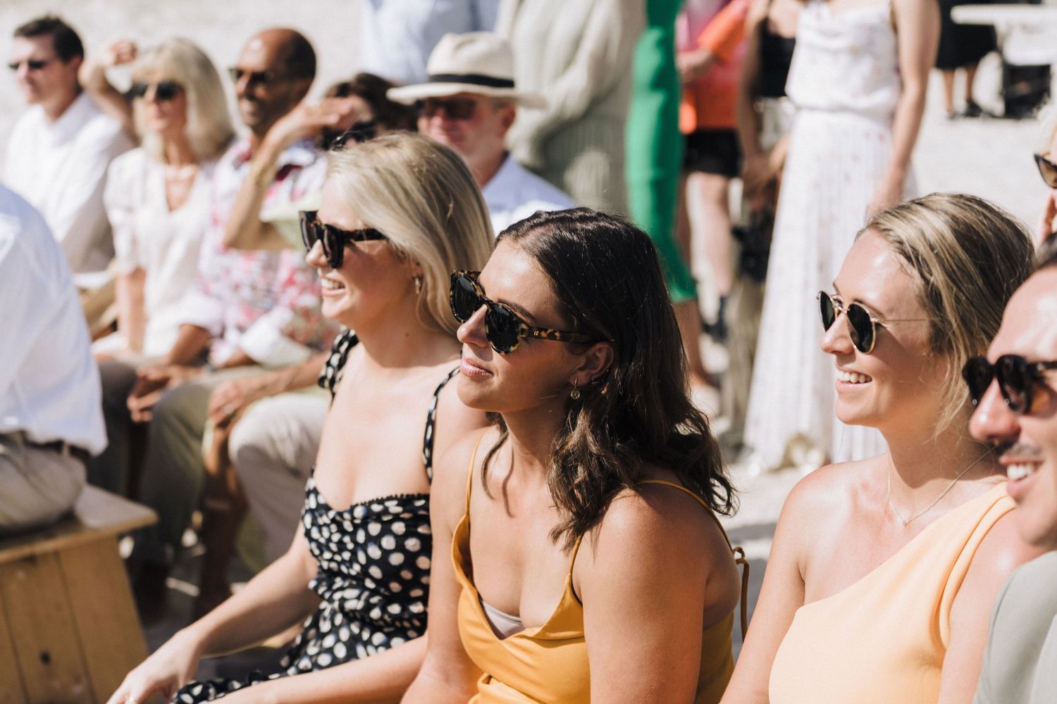 Wedding guests seated in outdoor rows wearing sunglasses and summer attire.
