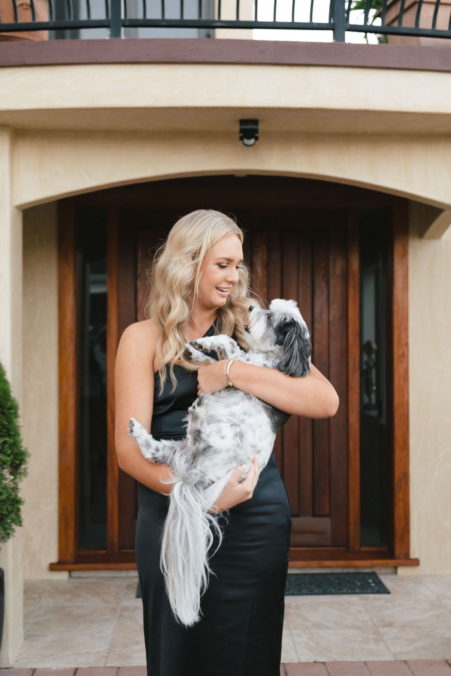 Woman in black dress smiling while holding and looking down at a white fluffy dog in front of wooden door.