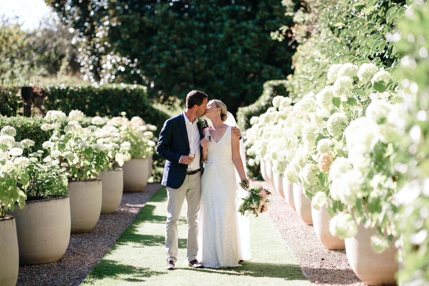 Couple sharing intimate moment along garden path lined with white potted flowers.