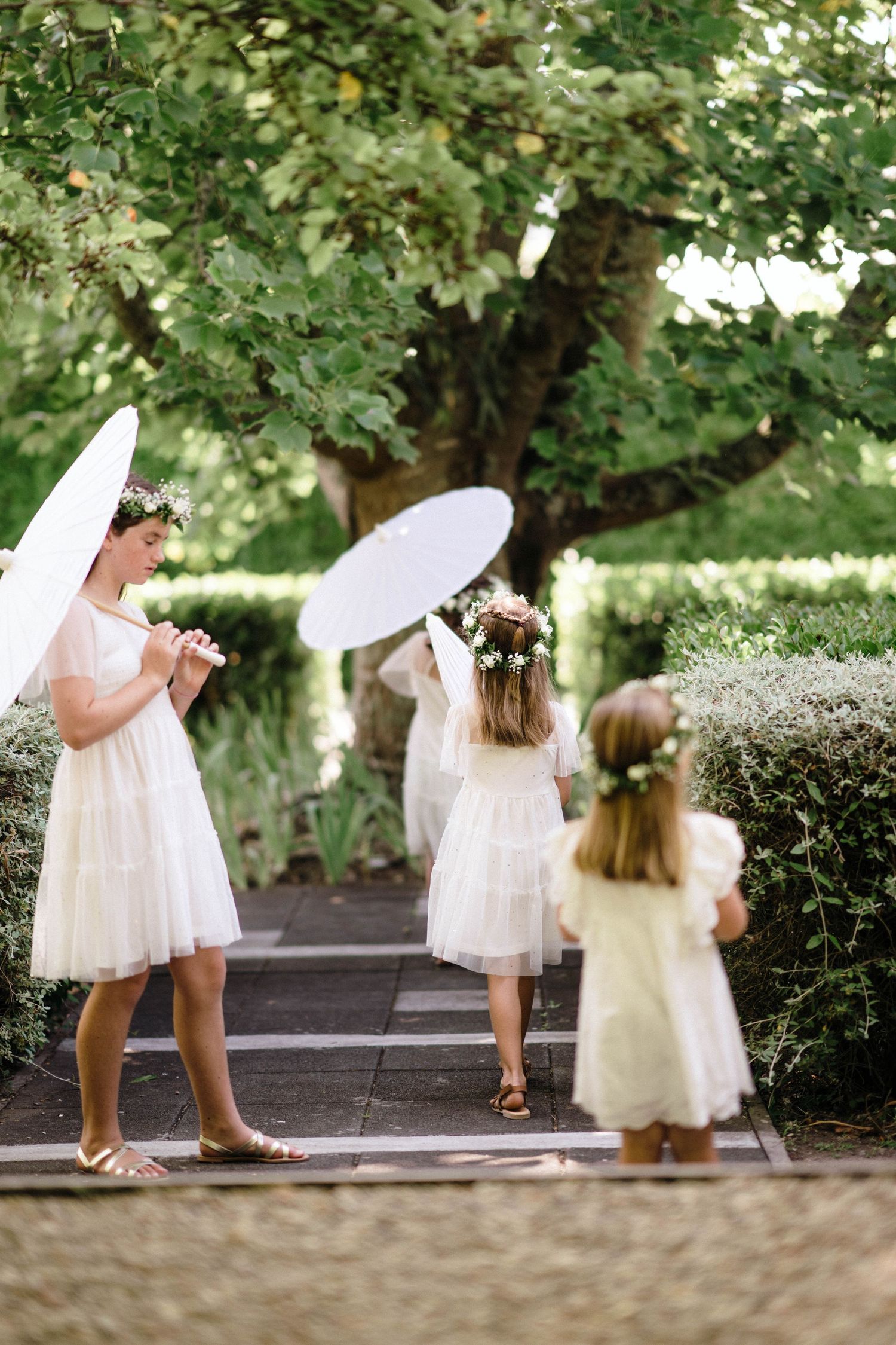 Three young attendants in white dresses holding parasols while standing on garden path.