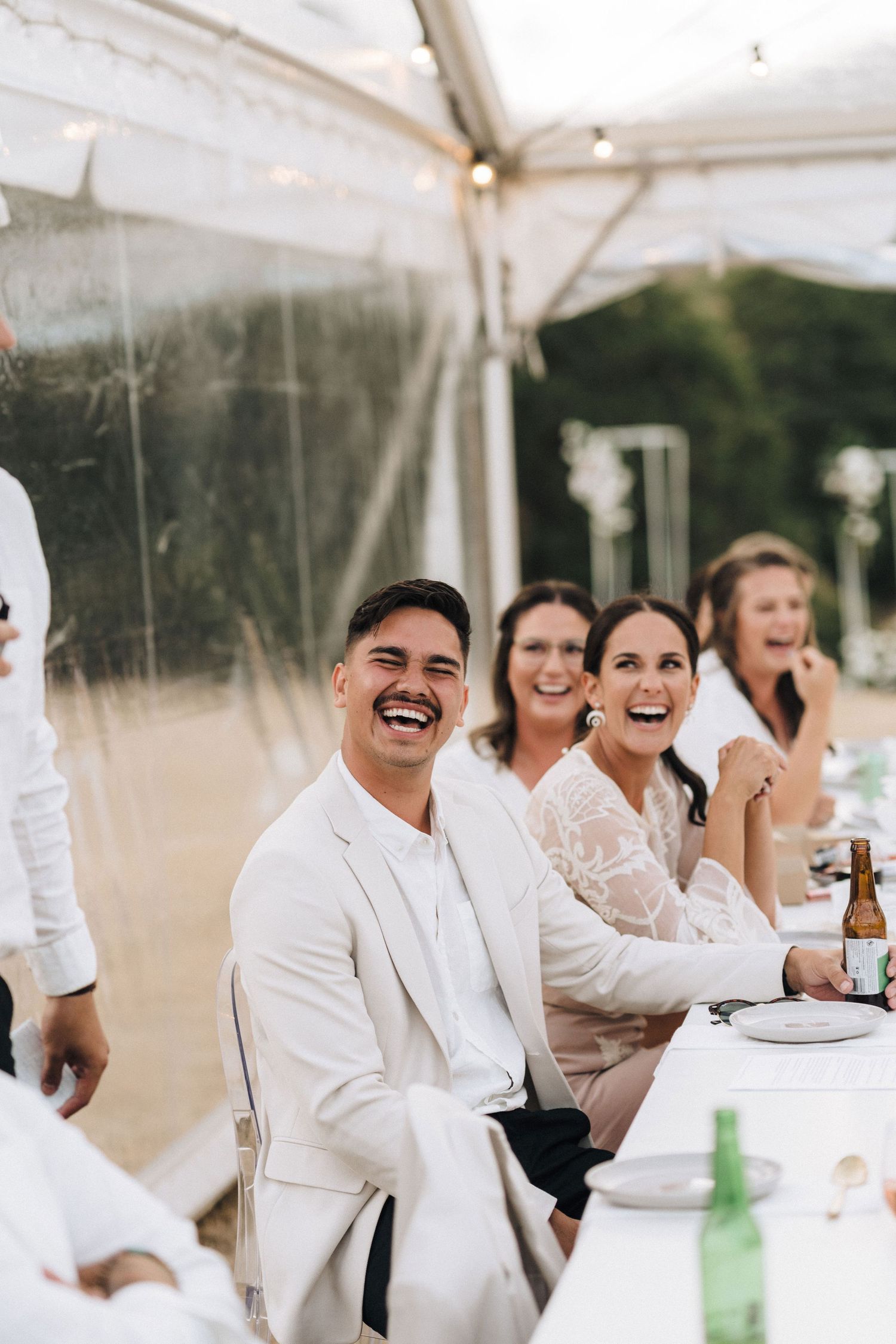 Wedding guests laughing and celebrating under a white tent at an outdoor reception.