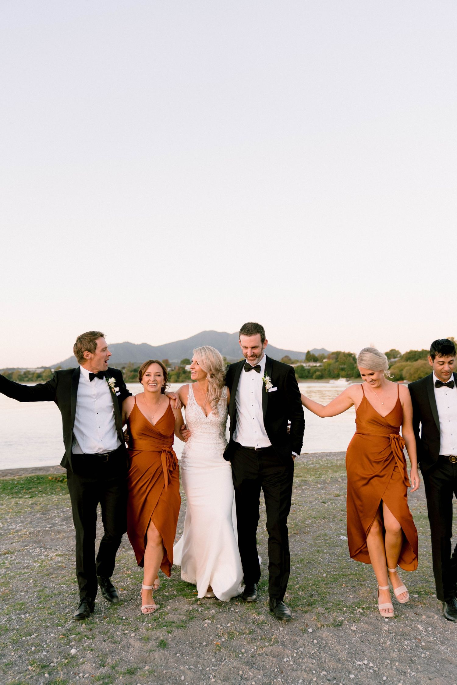 Wedding party in formal attire strikes playful poses together on a beach at sunset.