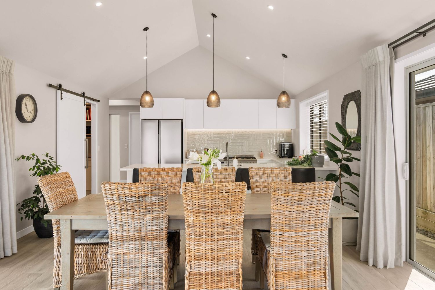 Modern dining room with rattan chairs, wooden table, and copper pendant lights beneath a vaulted ceiling.