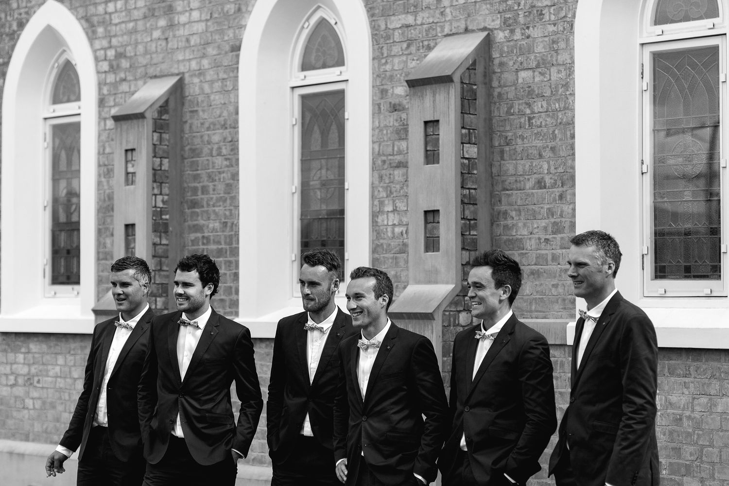 A row of men in dark suits stand against a brick building with arched windows in a black and white photograph.