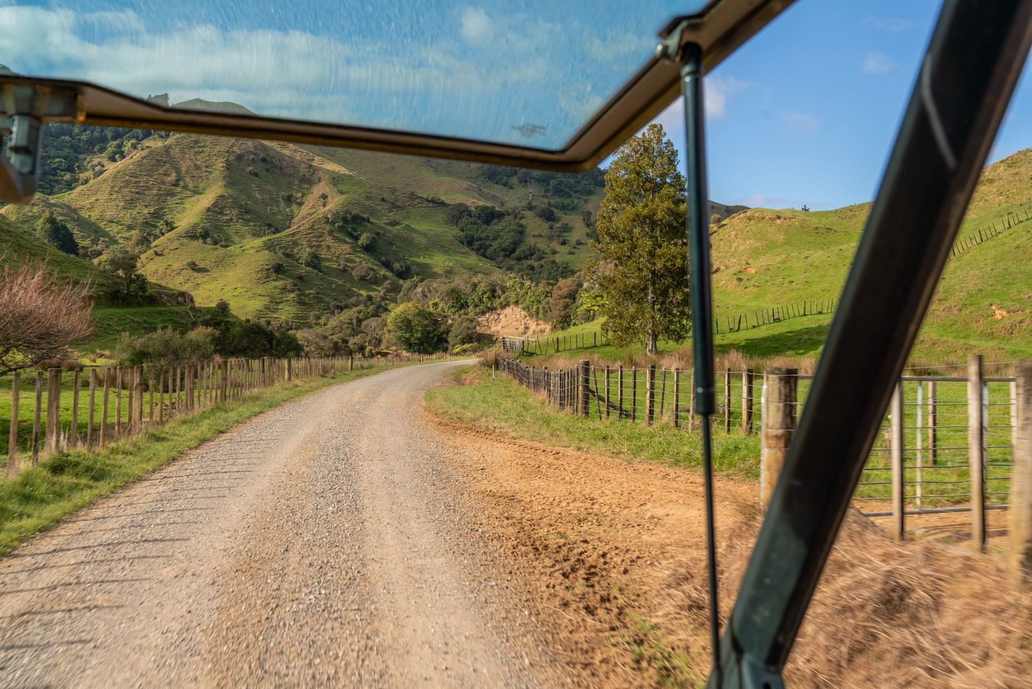Dirt road winding through green hills with wooden fence posts and stunning mountain backdrop.
