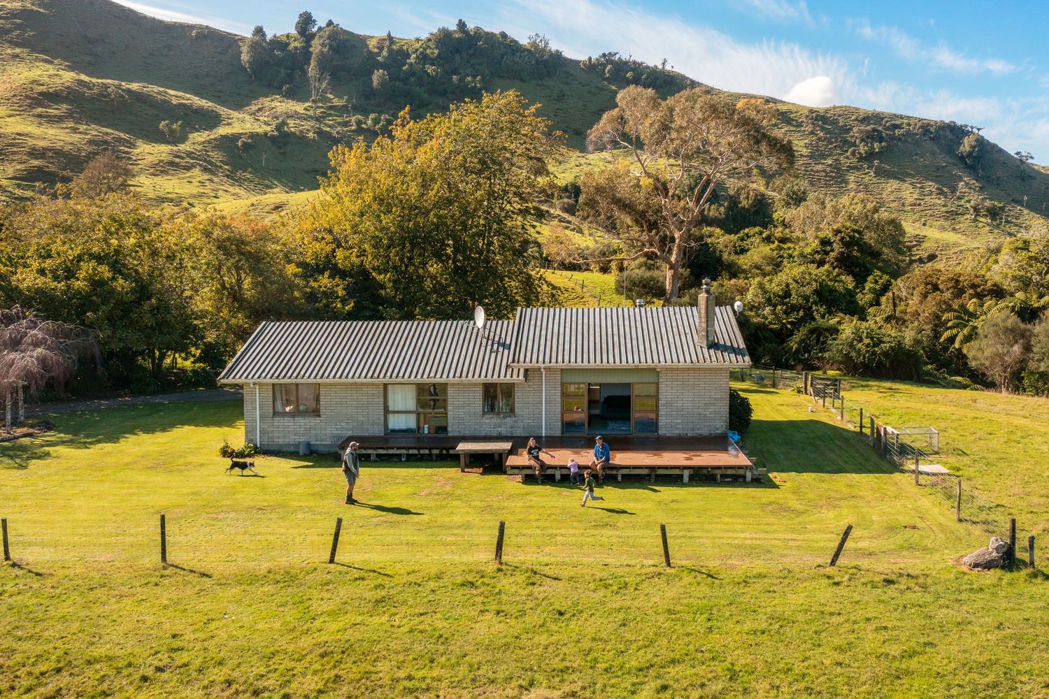 Rural farmhouse nestled in lush green hillside with wooden fencing and mountain backdrop.