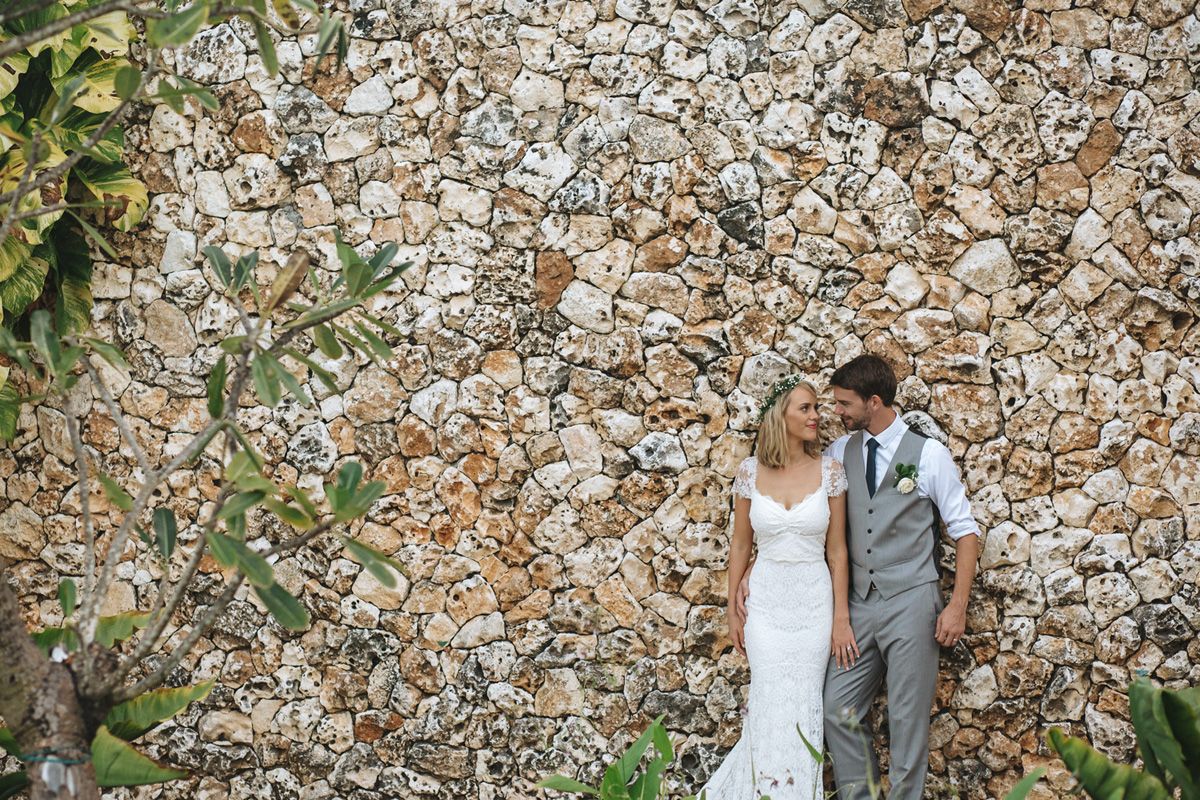 Bride and groom pose against a rustic stone wall with greenery.