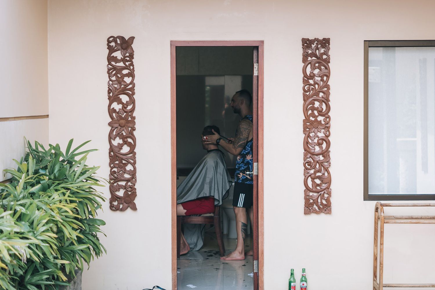 Wooden decorative panels flank white doorways along a beige building exterior with tropical plants and windows.